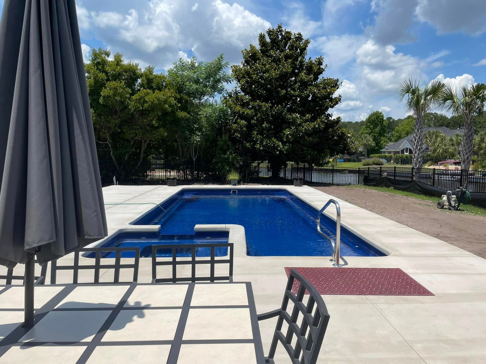 A rectangular blue swimming pool with concrete decking, patio furniture, and an umbrella, near a body of water and trees.