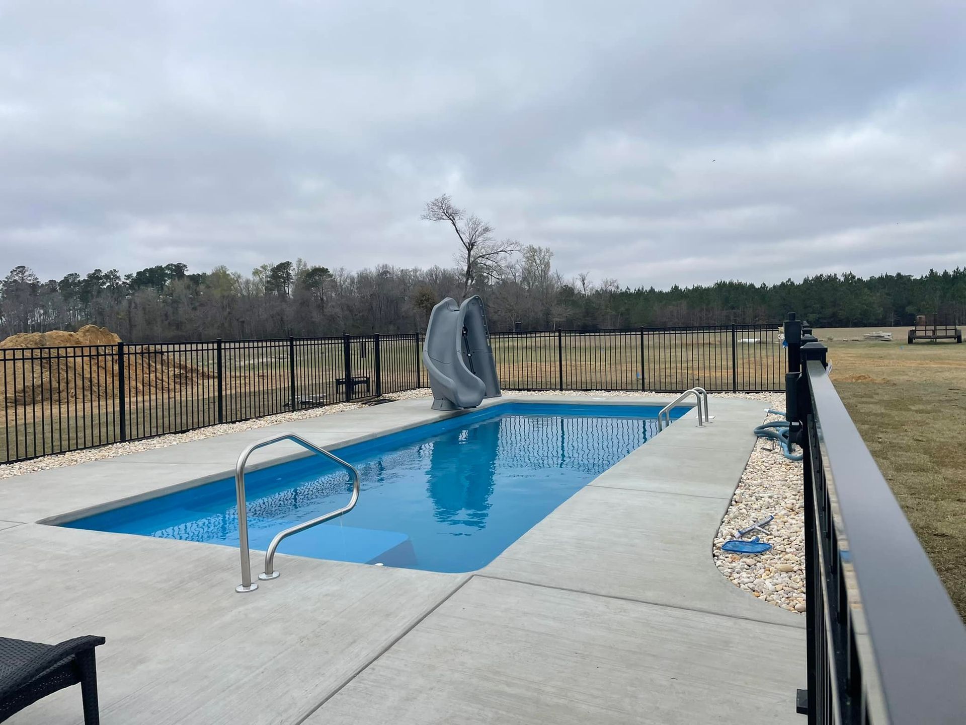 Rectangular outdoor pool with a slide, surrounded by a black fence and concrete patio. Cloudy sky.