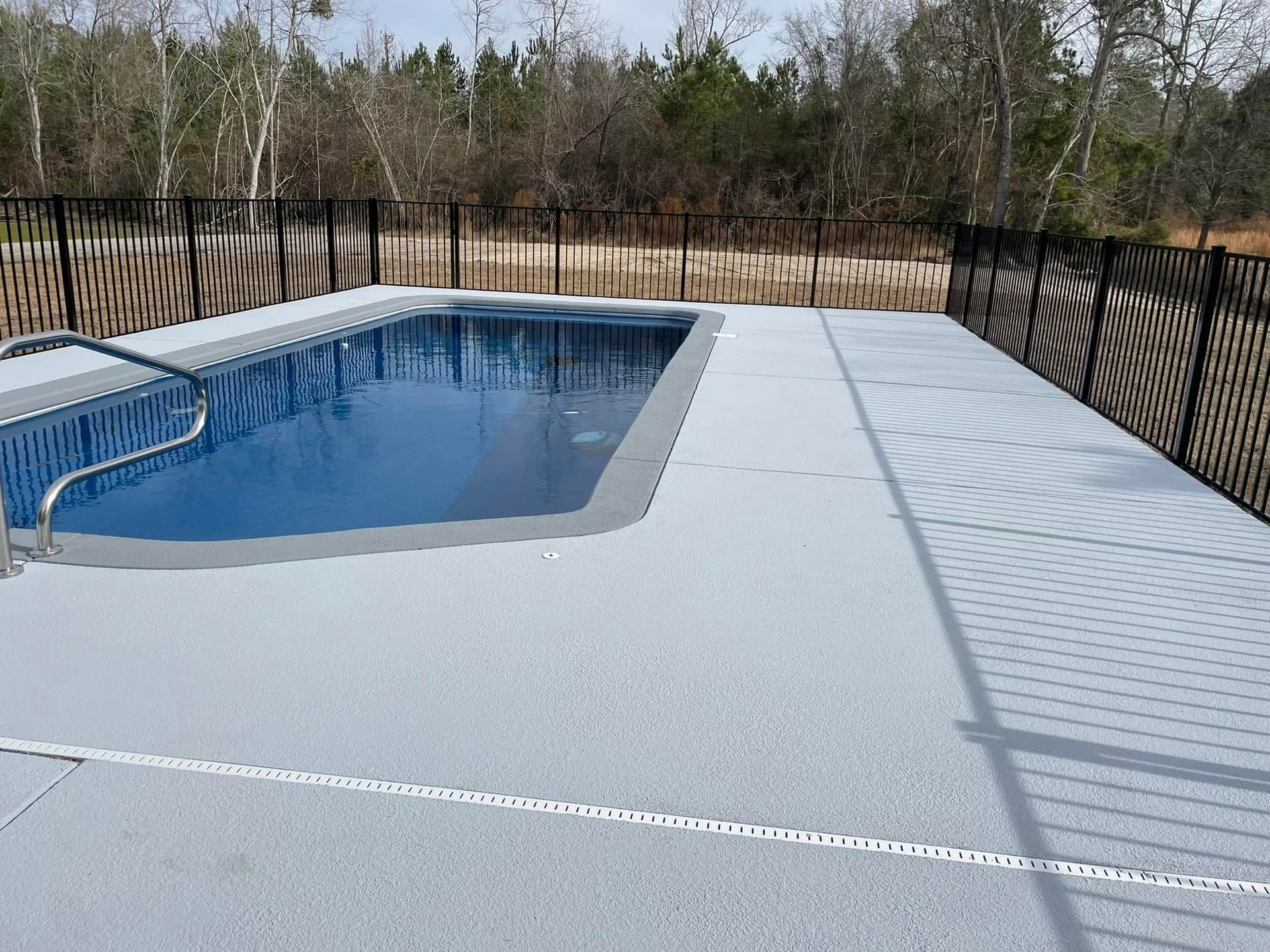 A rectangular pool with light gray concrete deck, black metal fence, and a wooded background.