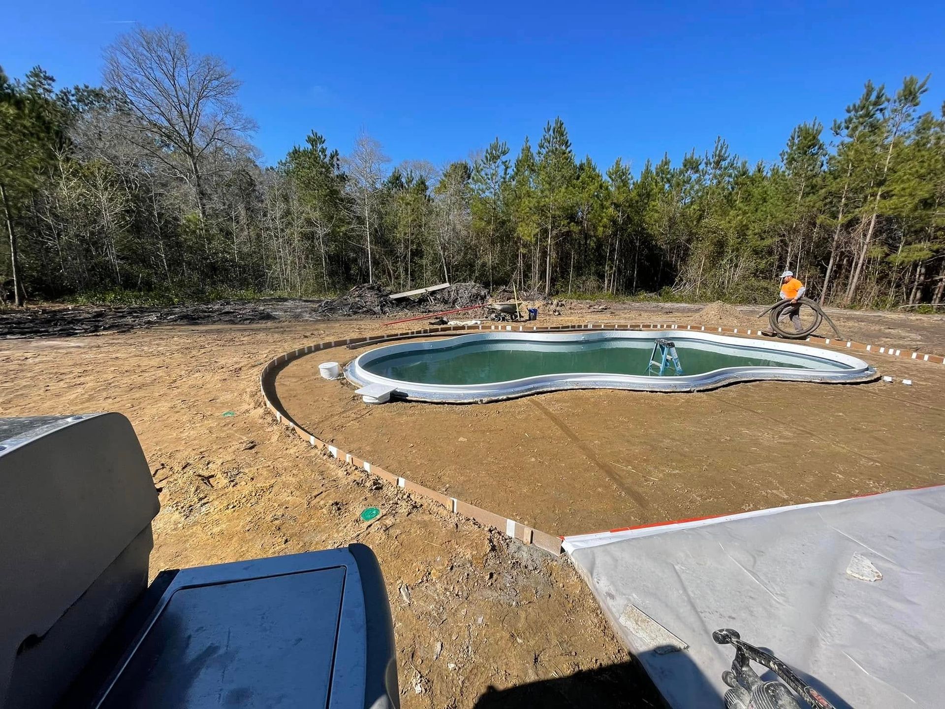 Pool under construction in a yard surrounded by trees. A worker is near the pool. Sunny day.