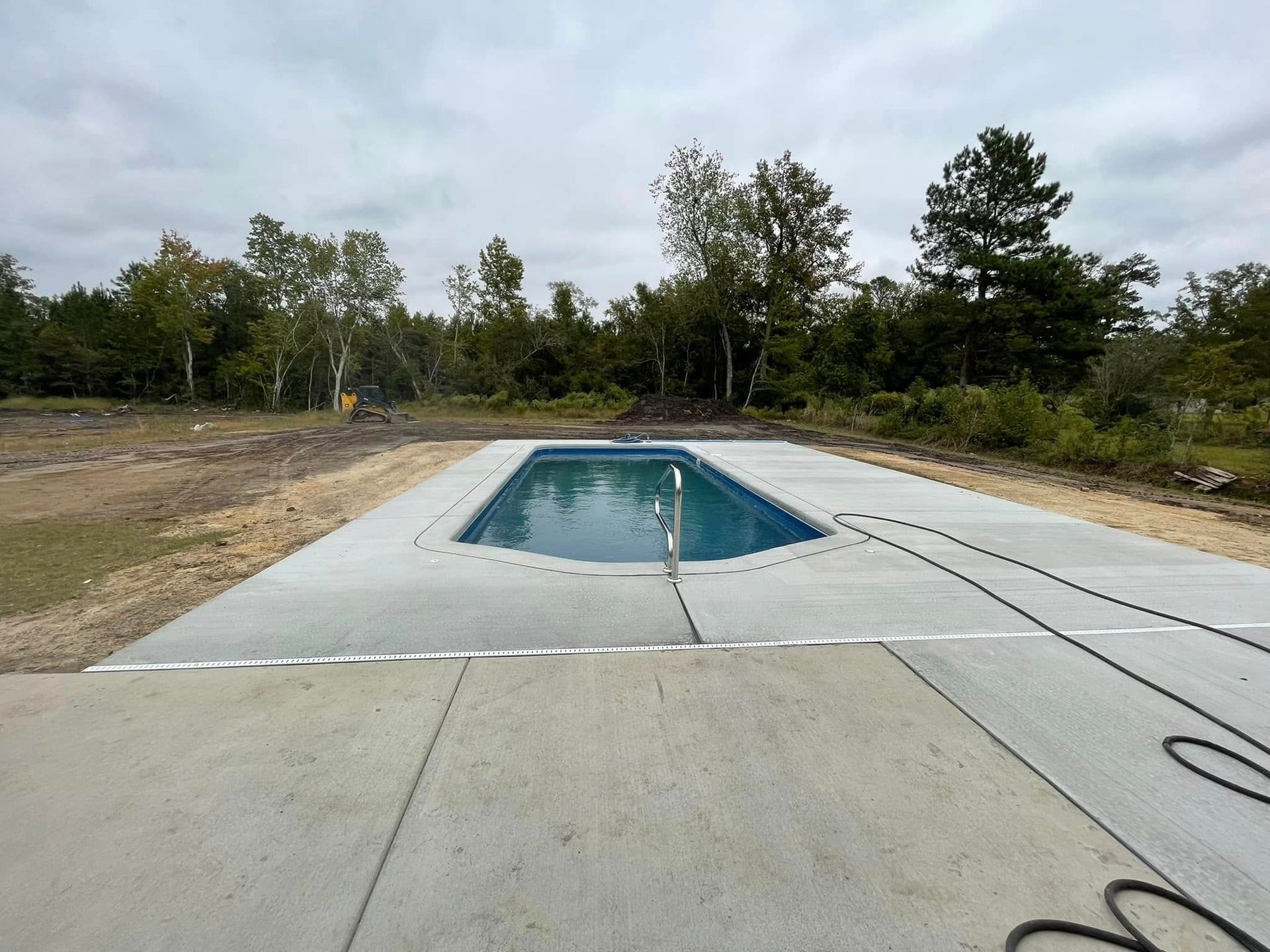 Concrete pool deck surrounding a rectangular pool filled with water, trees in the background, overcast sky.
