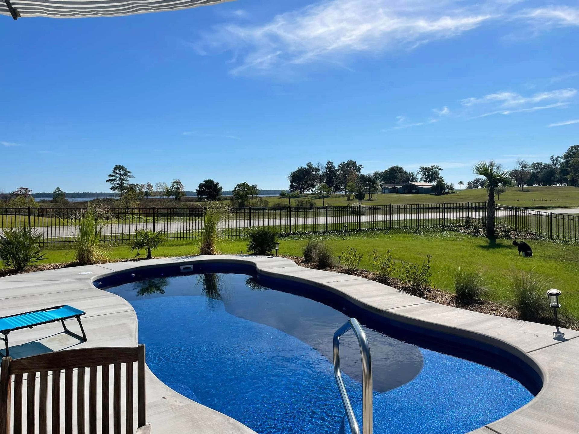 Pool with blue tile overlooking a grassy yard, fence, and water under a blue sky.