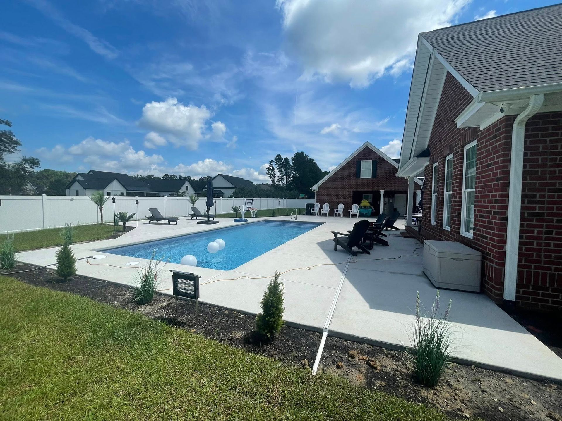 Backyard with a rectangular swimming pool, concrete patio, brick house, and white fence under a blue sky.