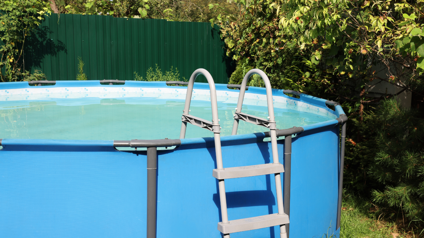 Blue above-ground pool with silver ladder, set against green fence and foliage.