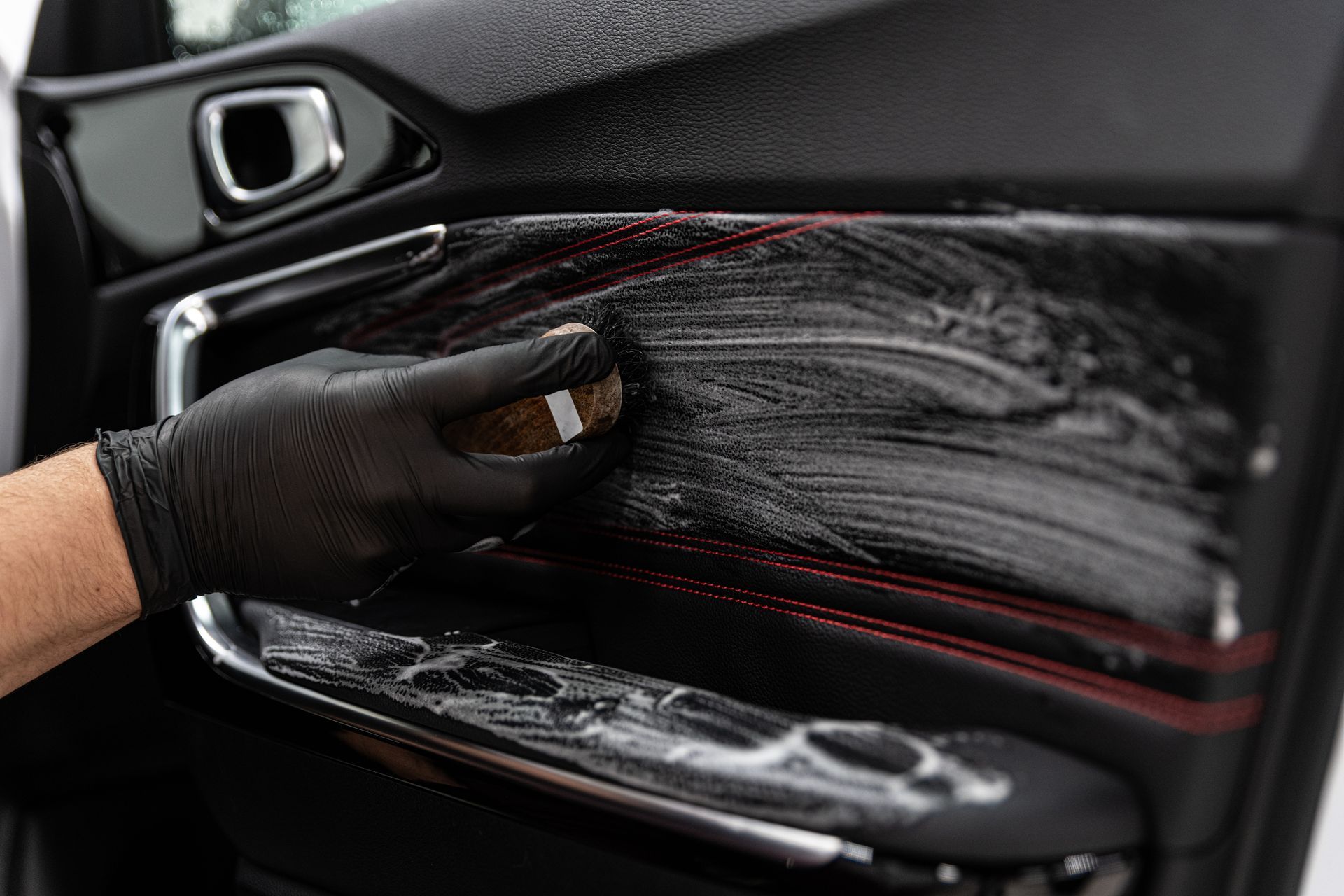 A gloved hand scrubbing a car door panel with cleaning foam during the interior detailing process