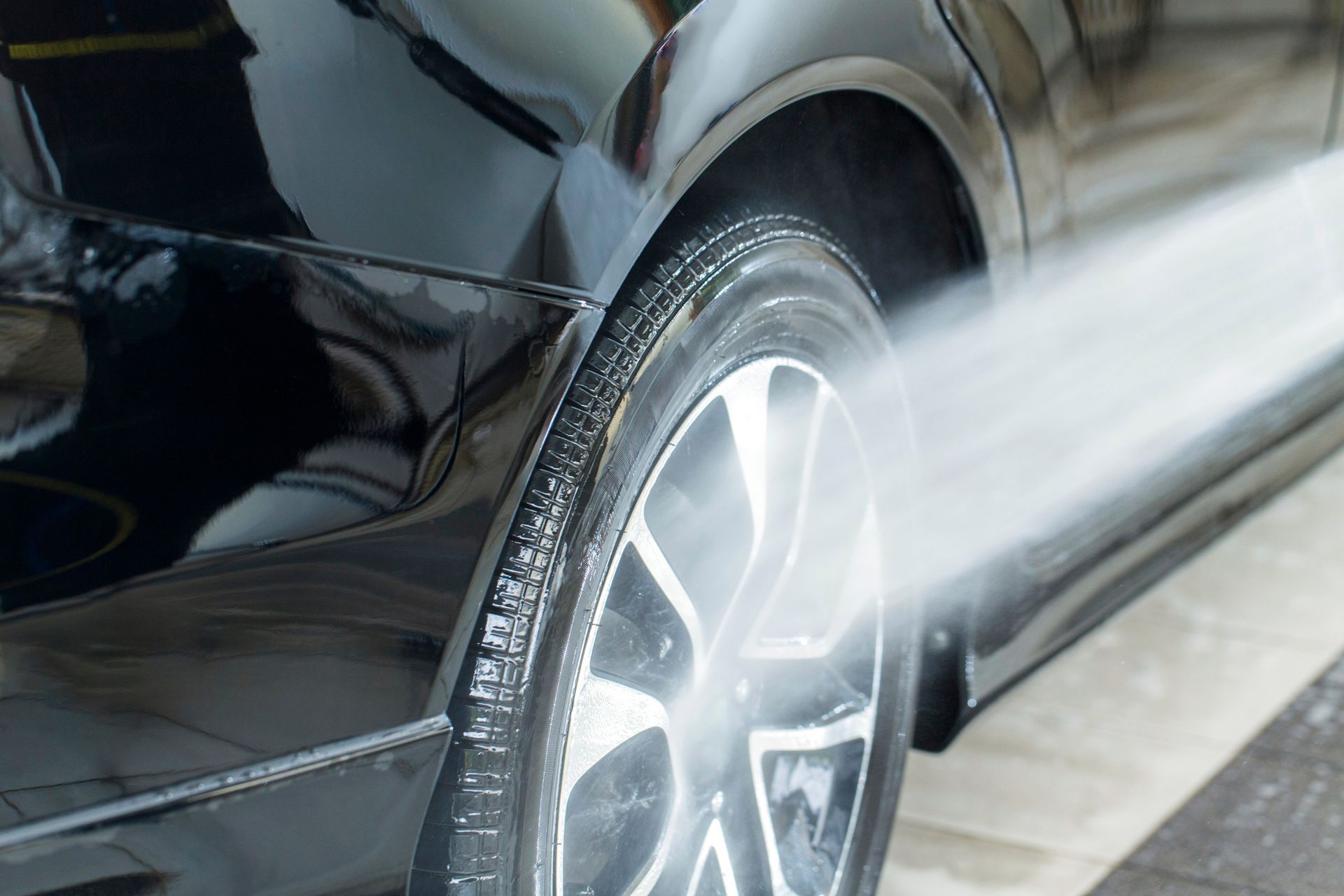 A high-pressure water jet cleans a shiny black car’s tire during a detailing service.
