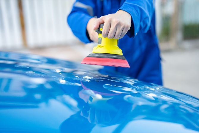Person in blue overalls using a yellow electric polisher on a blue car.