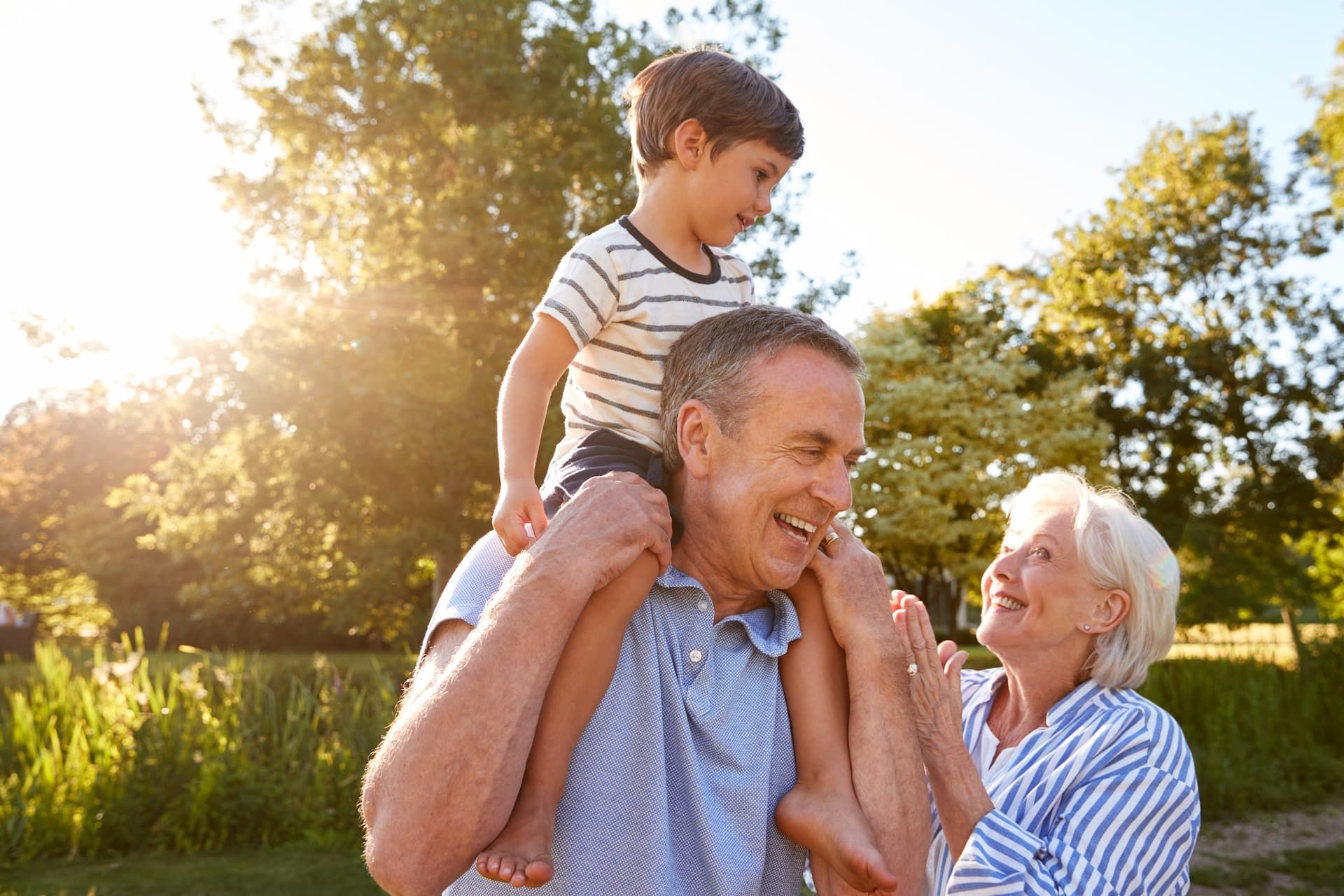 Smiling man carrying a child on his shoulders while a woman smiles nearby in a sunlit park