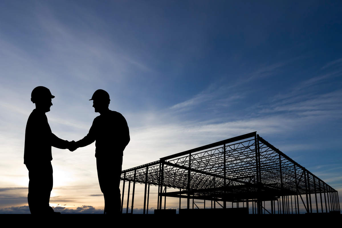 Silhouetted construction workers shaking hands in front of a building frame under a blue sky.
