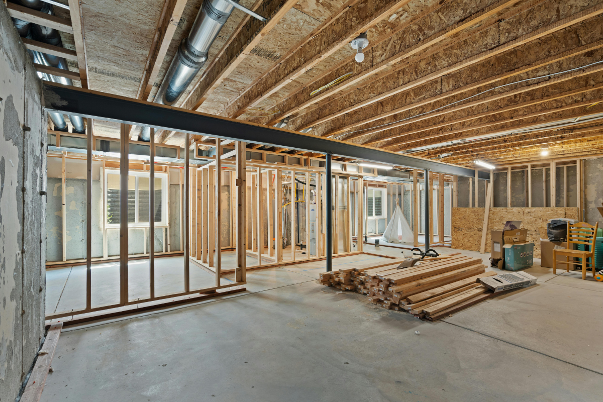 Basement under construction, wood framing and beams. Cement floor, visible ceiling rafters, and materials.