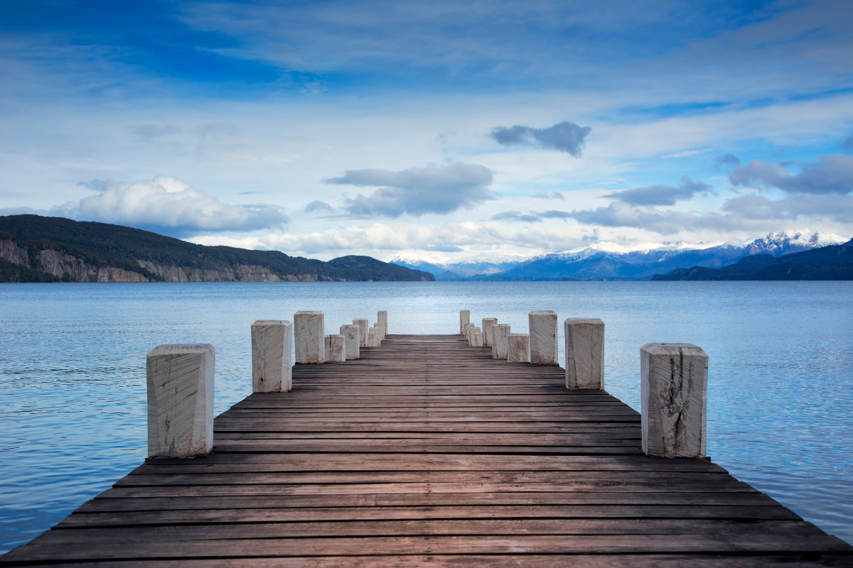 Wooden pier extending over calm blue water towards mountains under a cloudy sky.