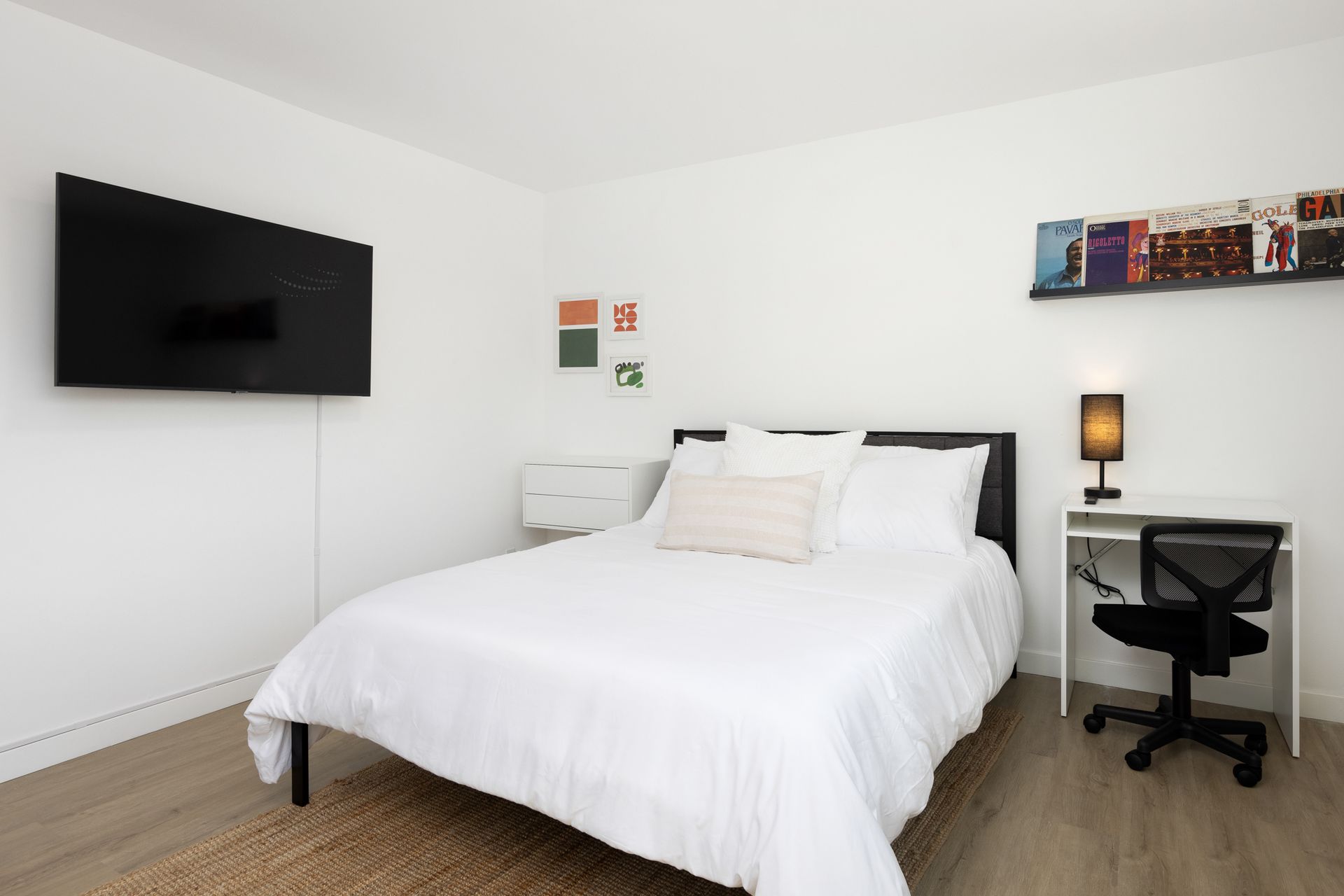 A minimalist bedroom with a bed, wall-mounted TV, small white desk with a chair, and a bookshelf on a white wall.