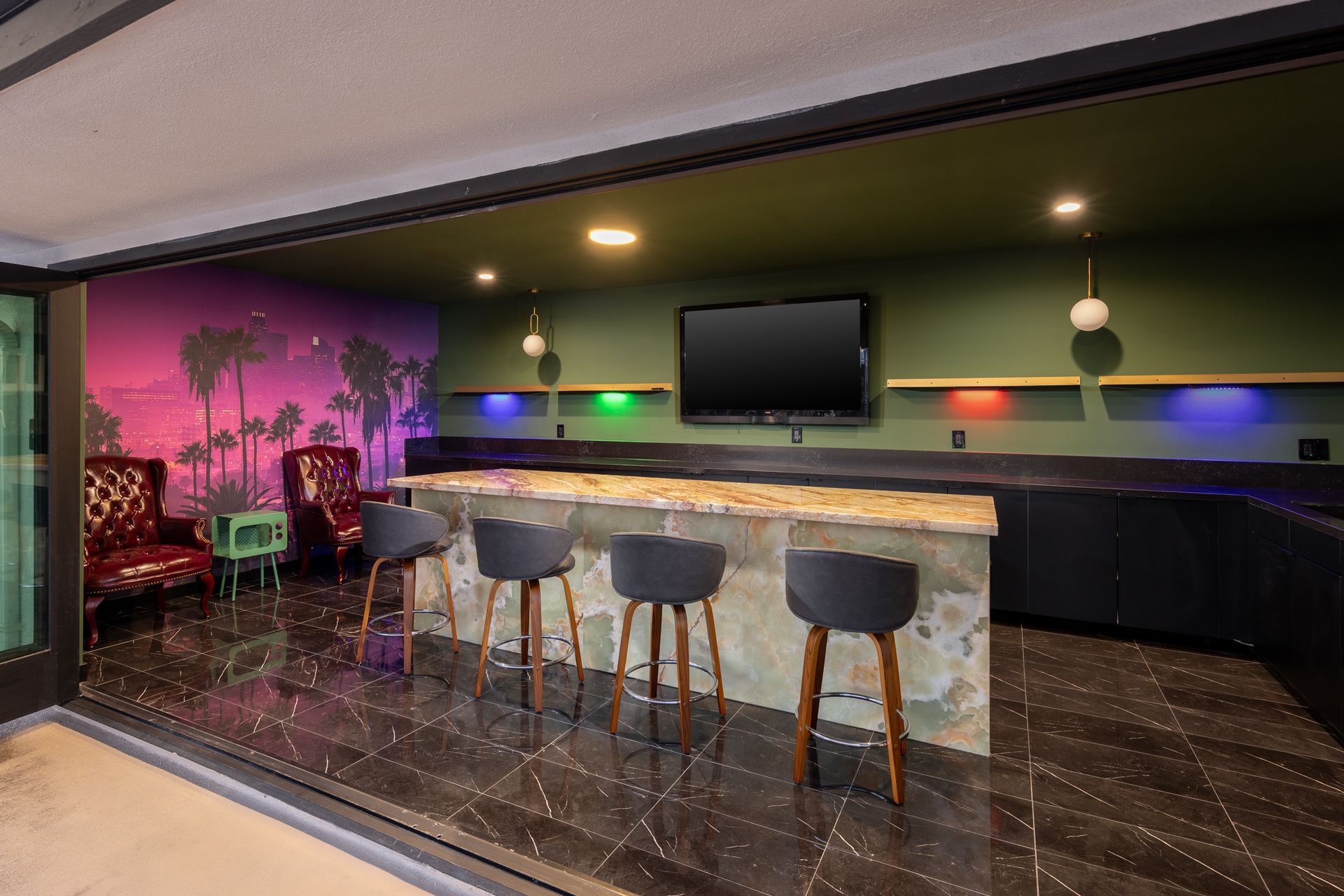 A modern home bar with stone counters, four dark stools, two red armchairs, neon palm art, and colorful under-shelf lights.