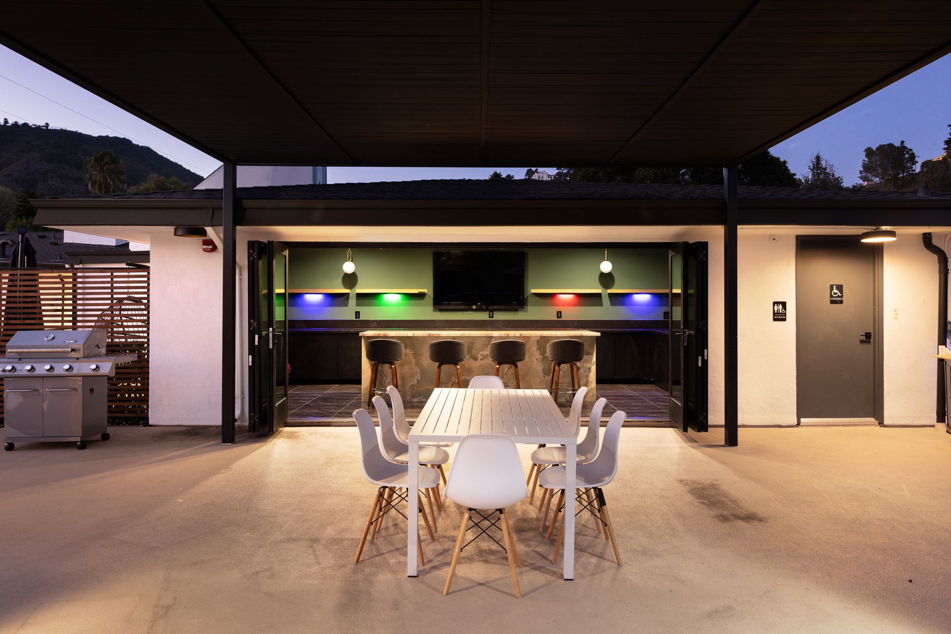 A patio area with a white dining set and outdoor kitchen with a bar, colorful LED lighting, and a grill at dusk.