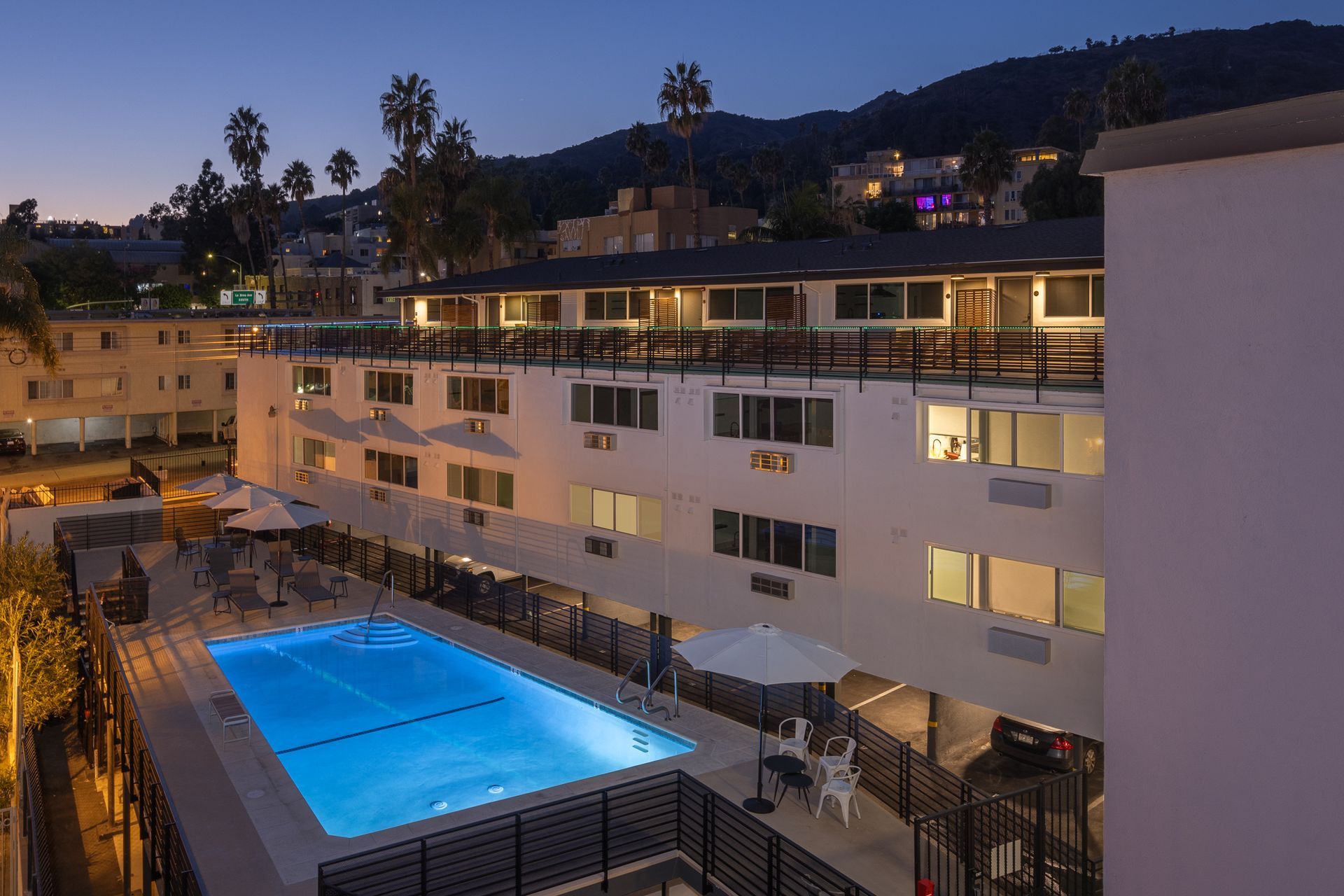 An outdoor swimming pool at a multi-story apartment complex at dusk, with lit windows and palm trees against a dark sky.