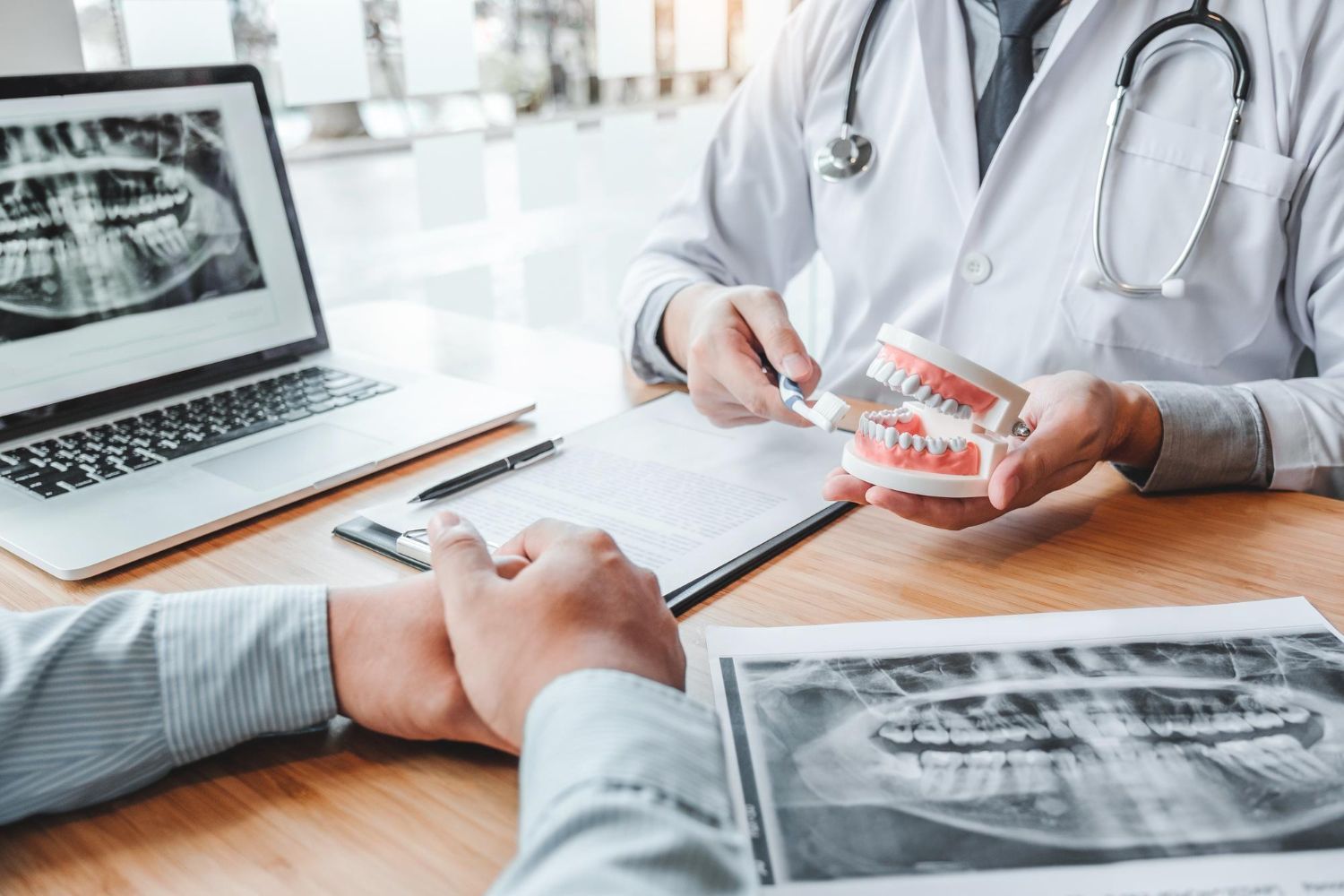 Dentist showing dental model and X-rays to a patient. Office setting with laptop and clipboard.
