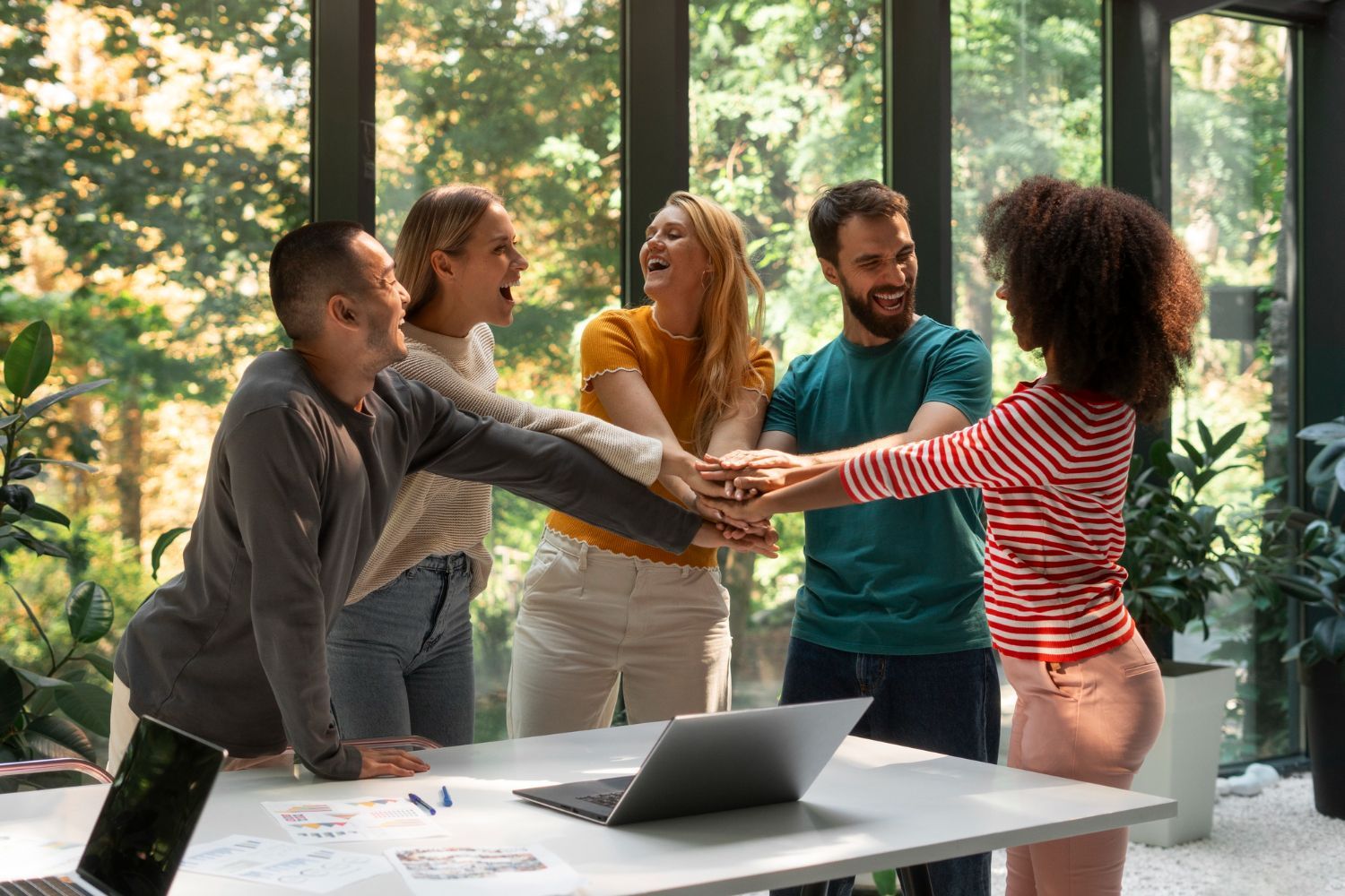 Five people huddle with hands together over a desk, laughing, in an office with large windows.