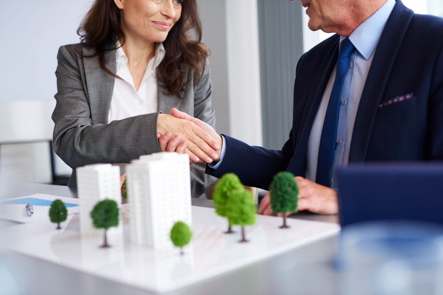 Woman and man shaking hands over a model of buildings with trees.