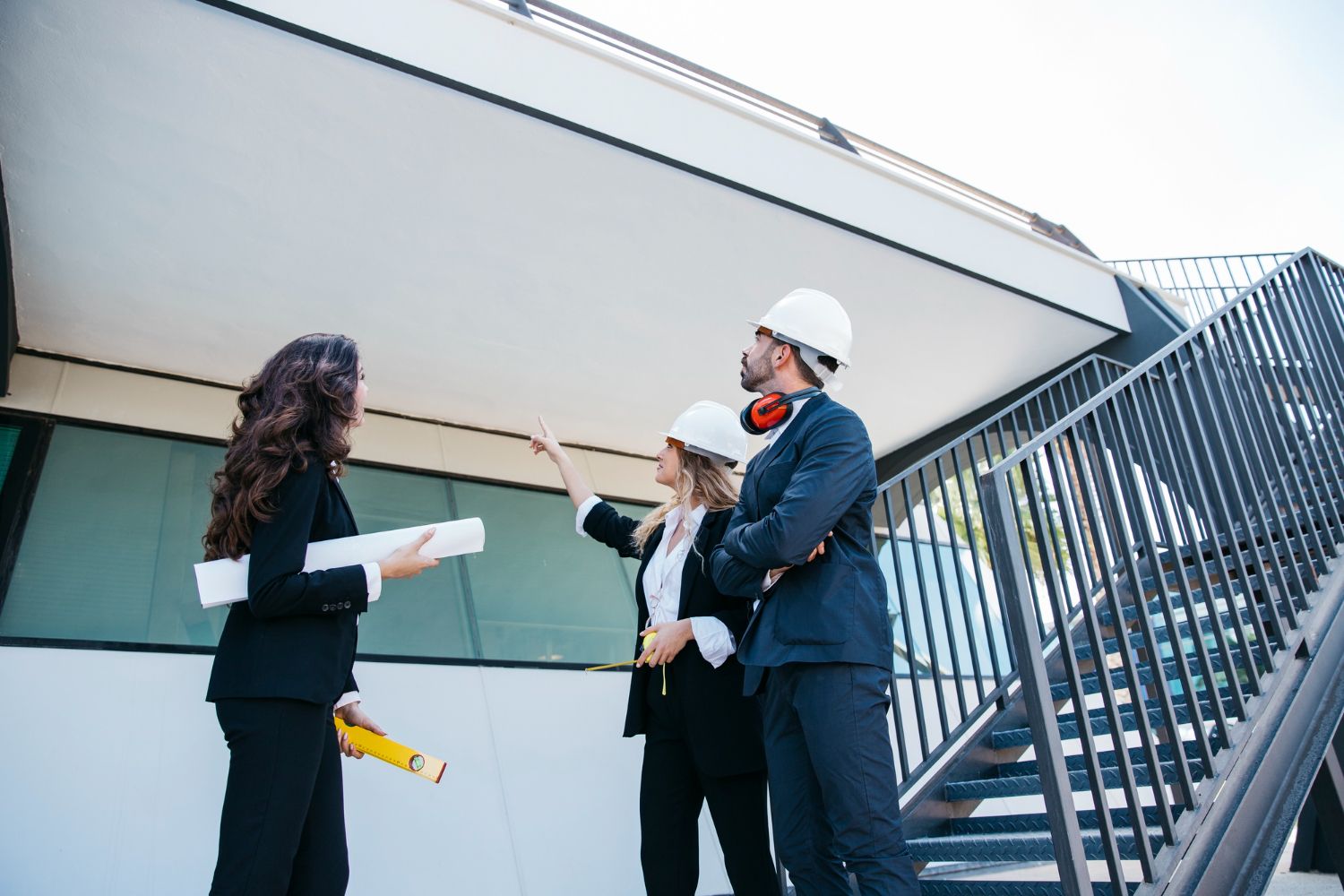 Three people in business attire and hard hats inspect a building exterior.