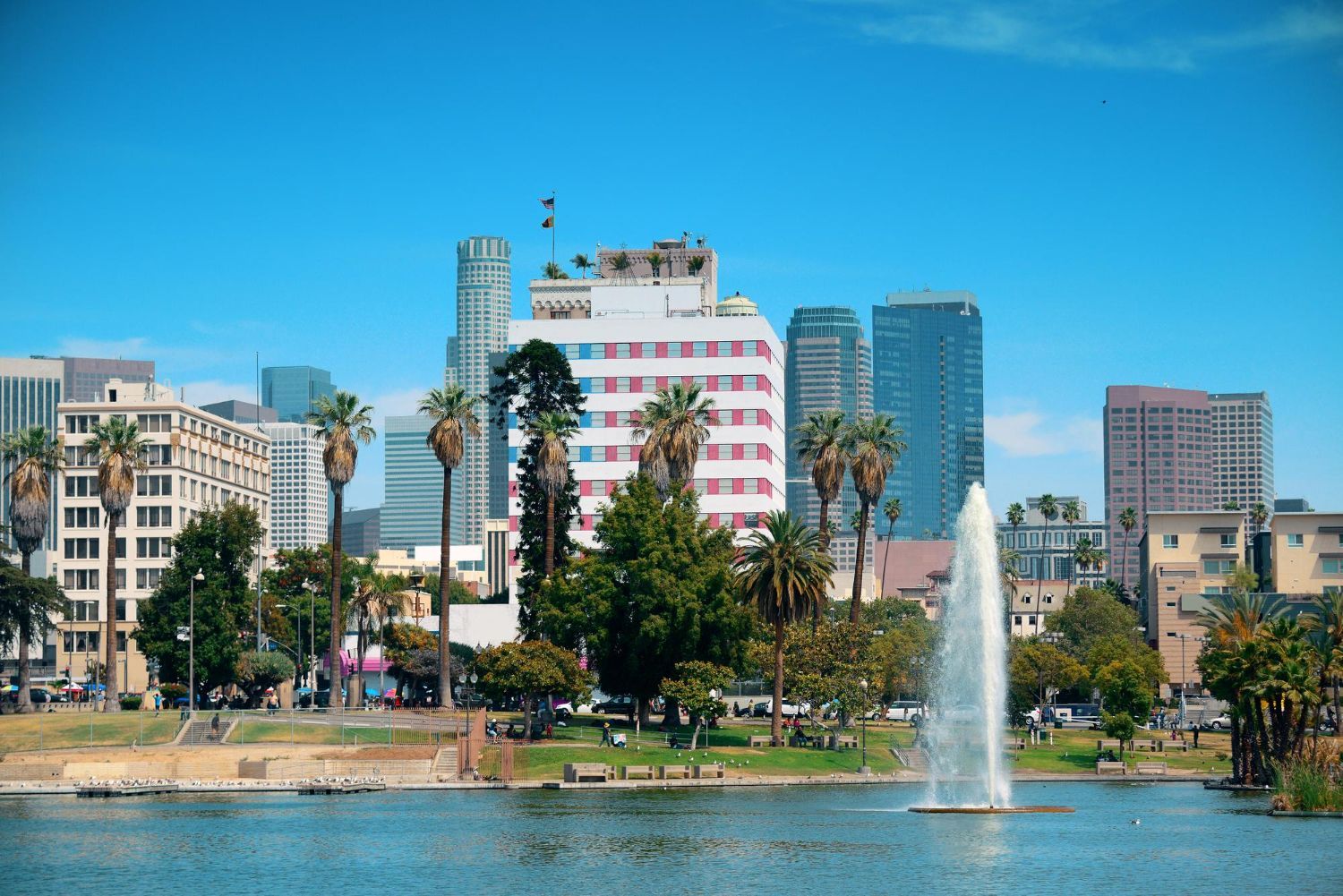 Downtown Los Angeles skyline with palm trees, a lake, and fountain against a blue sky.