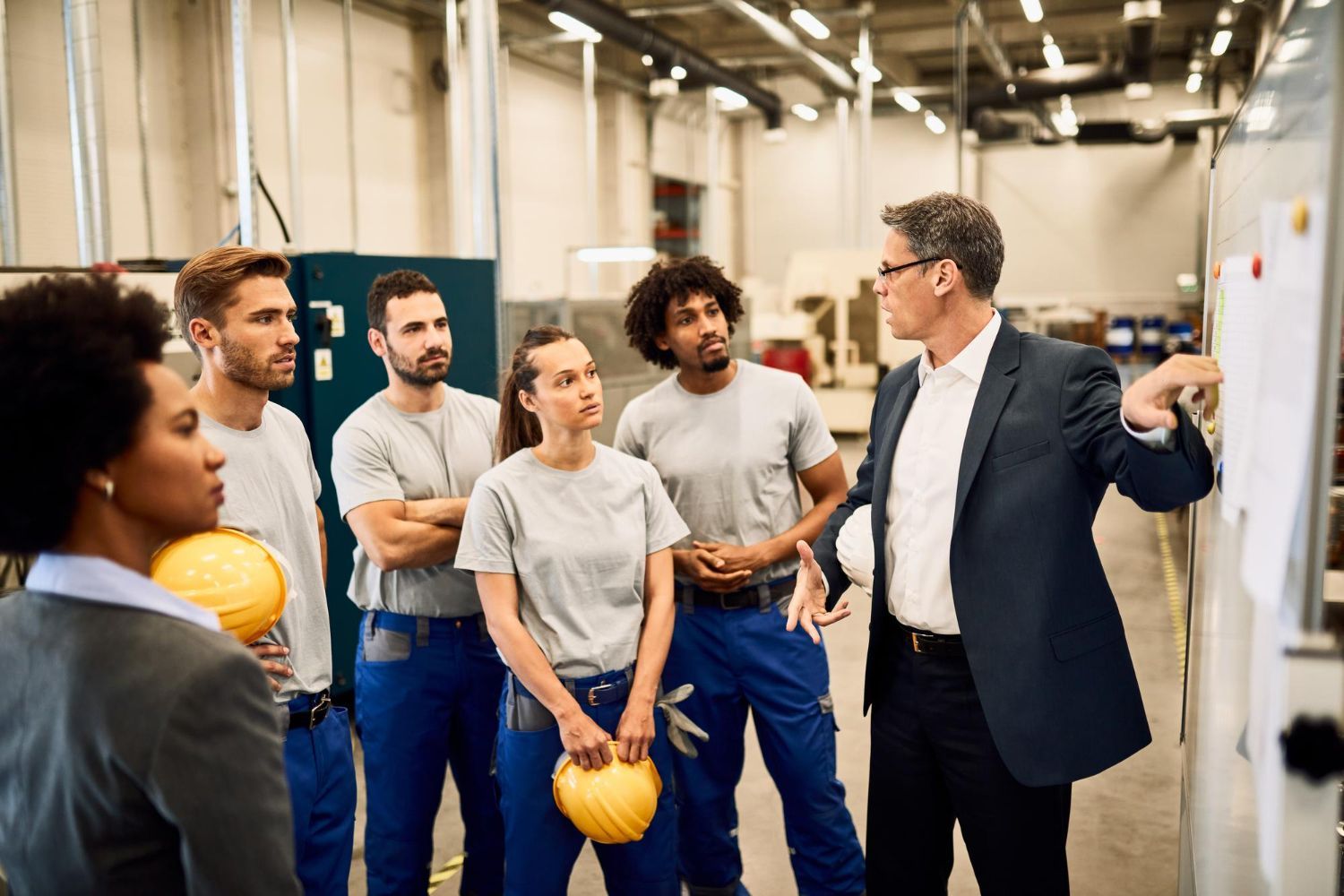 Group of workers in a factory setting, listening to a man in a suit pointing at a whiteboard.