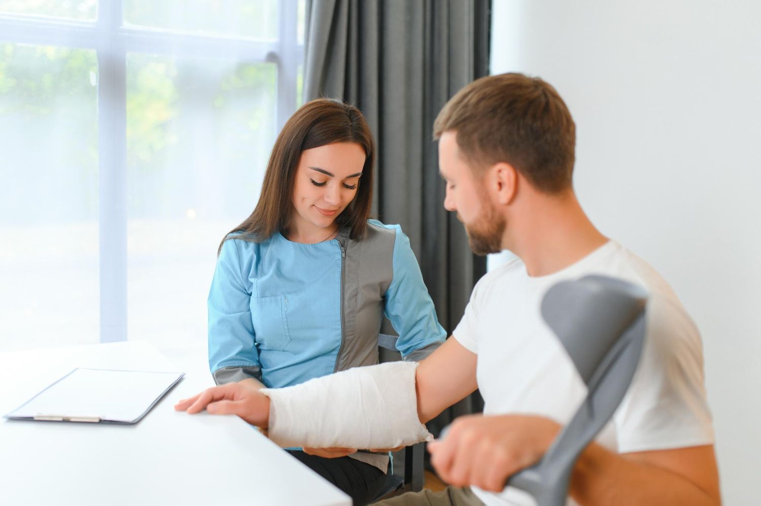 Woman assisting a man with an arm cast, crutch visible. Light-colored room, they are seated.