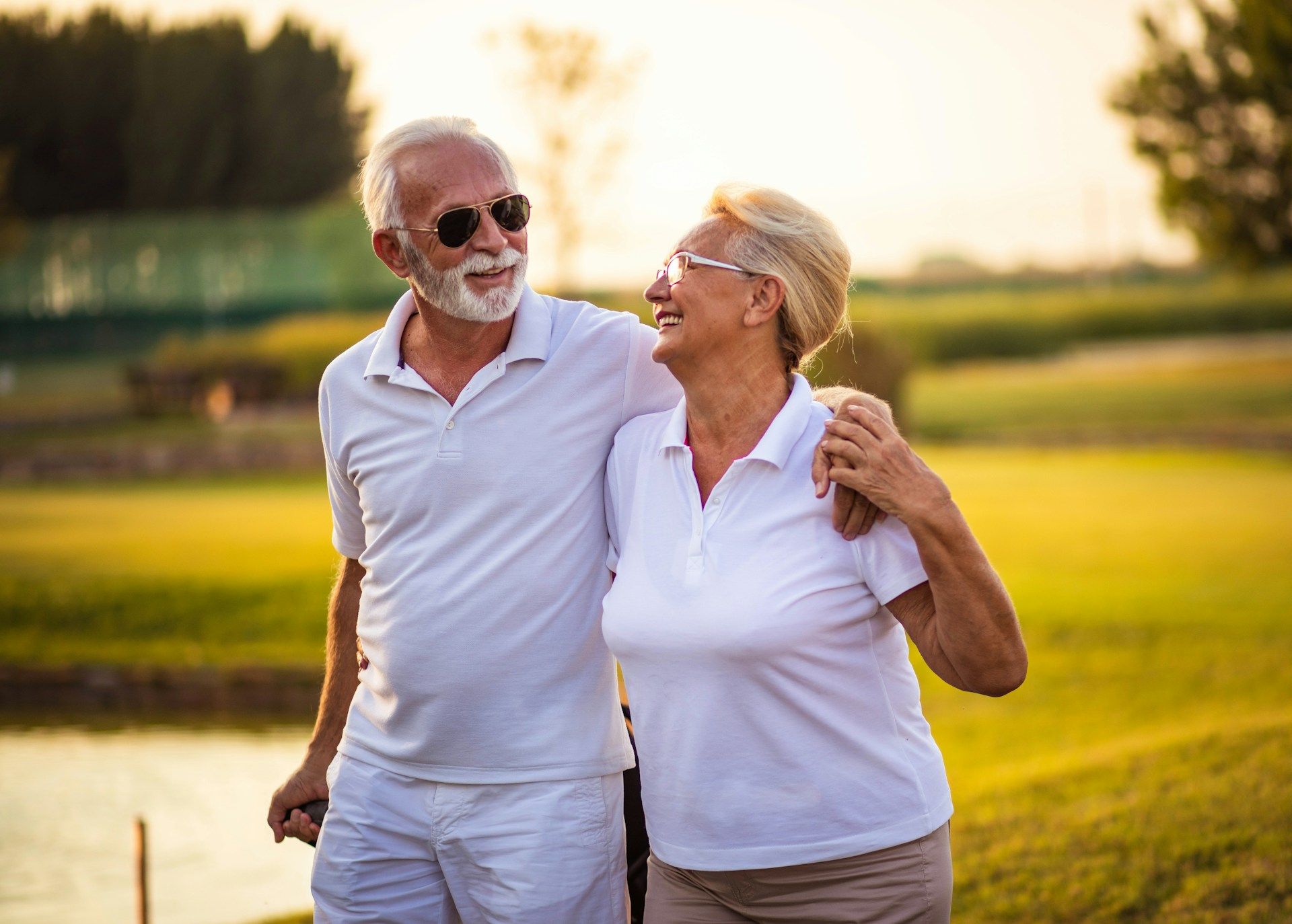Couple wearing white shirts, smiling, walking on a golf course. Man has sunglasses, arm around woman.