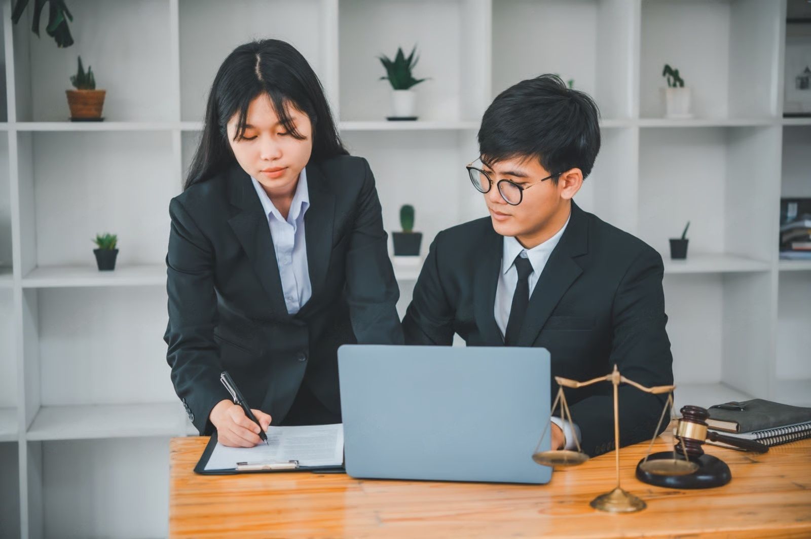 Two people in suits working at a desk with a laptop and documents. A gavel and scales are present.