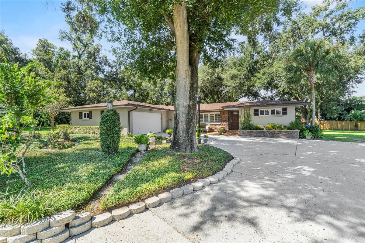 Ranch-style house with gray siding, driveway, and lush landscaping; tall trees in the background.