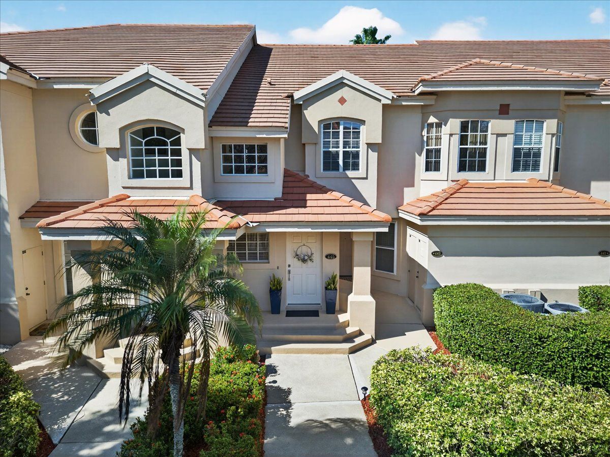 Two-story townhouse with a tan stucco facade, red tile roof, and a small front yard with palm trees.