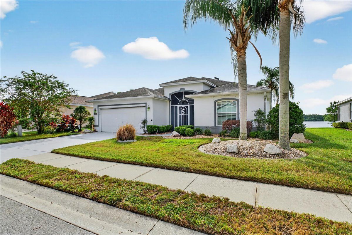 A light-colored house with a garage, front door, and palm trees in a grassy yard under a blue sky.