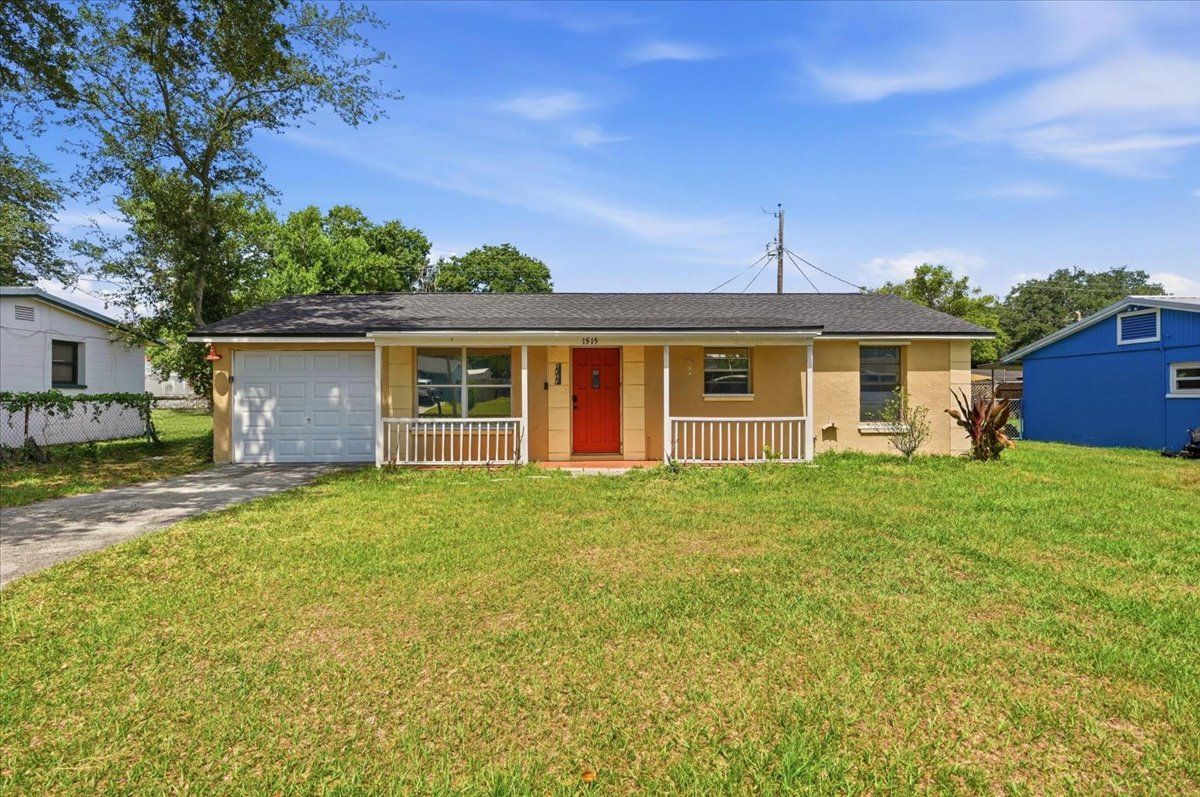 Single-story house with tan siding, red door, and a wide green lawn under a clear blue sky