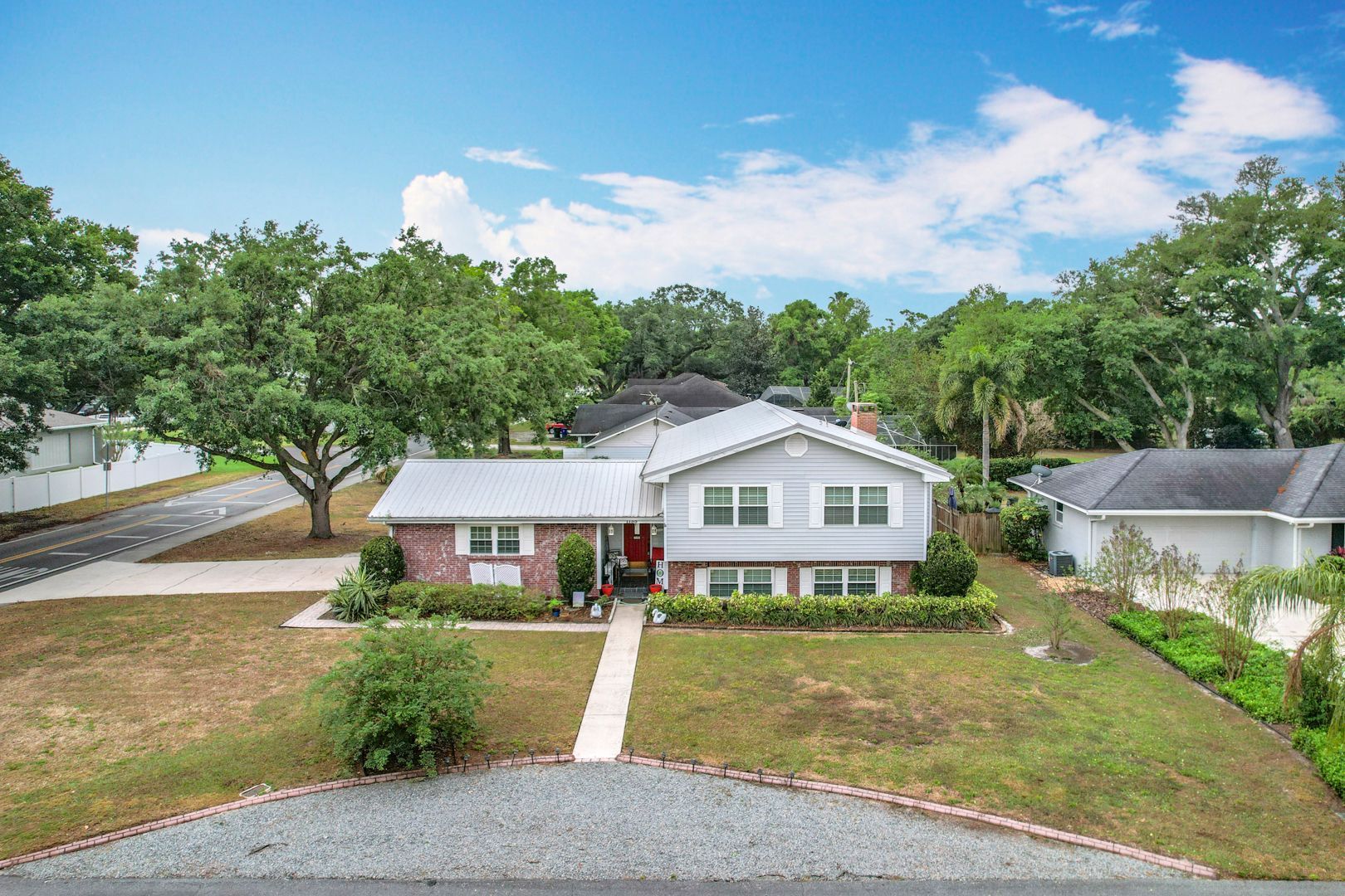 Two-story house with gray siding, brick facade, and a metal roof; surrounded by trees and a green lawn.