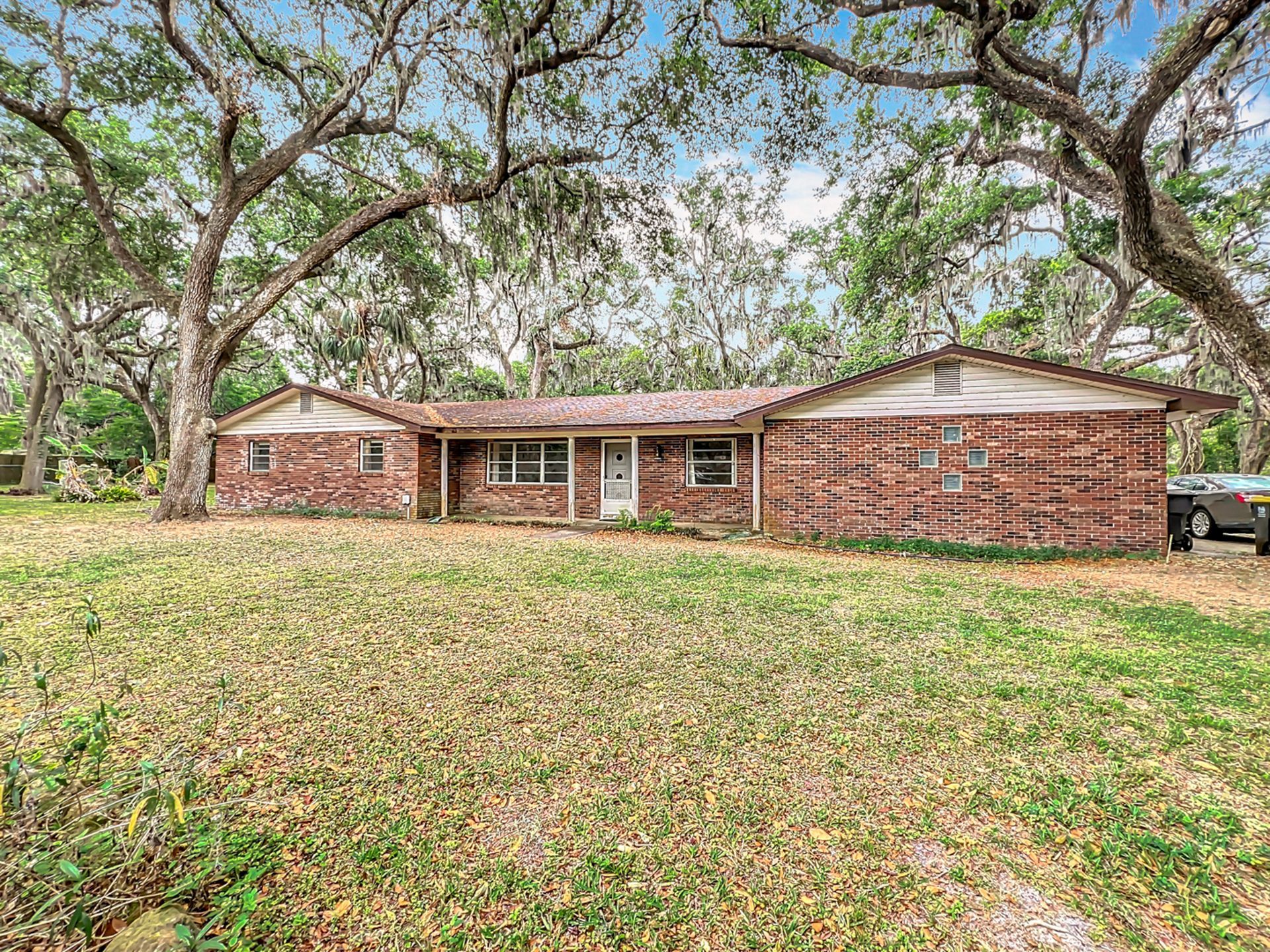 Brick ranch-style house nestled under large trees in a grassy yard.