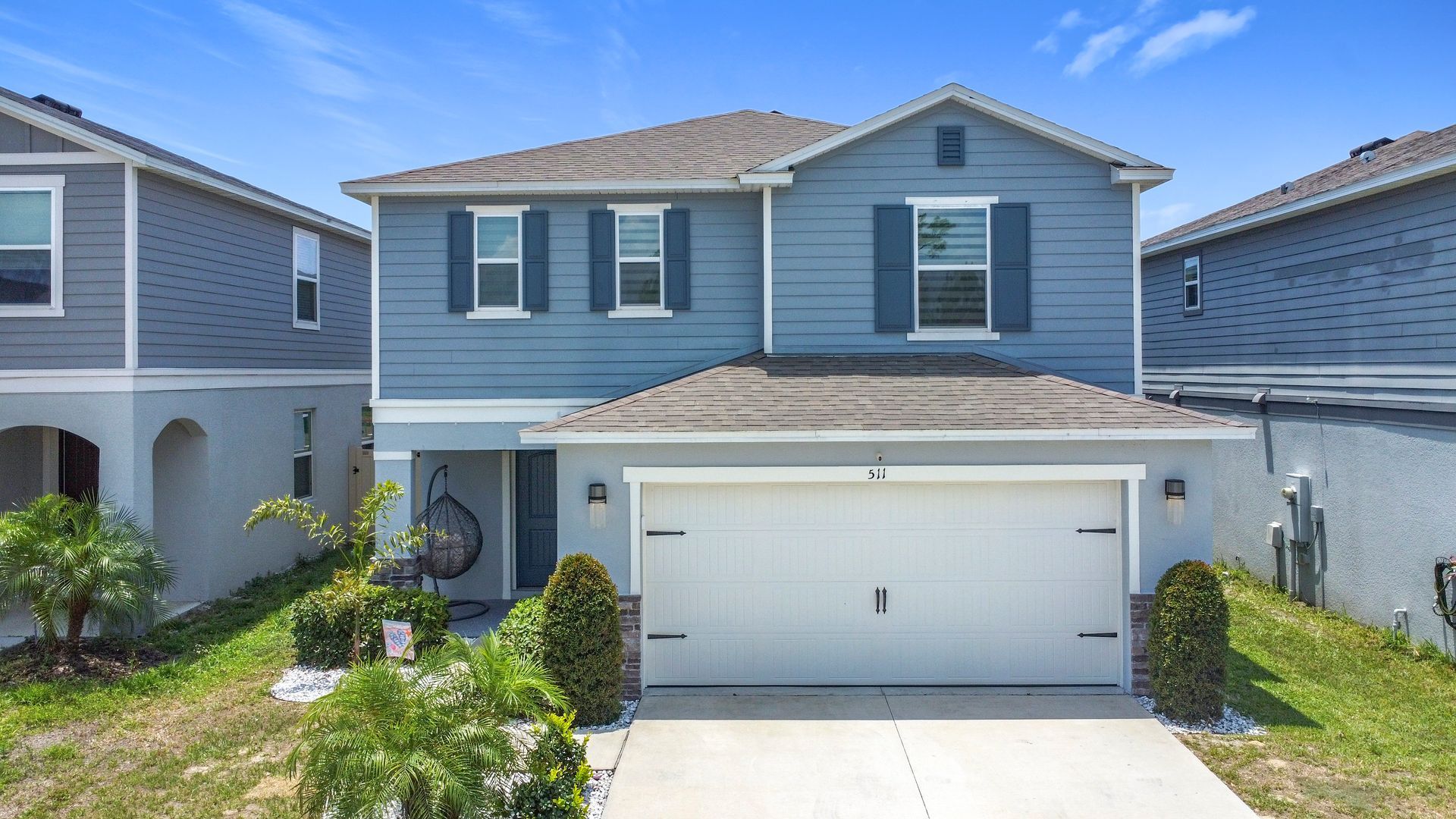 Two-story blue house with white garage door and shutters, in a residential neighborhood on a sunny day.