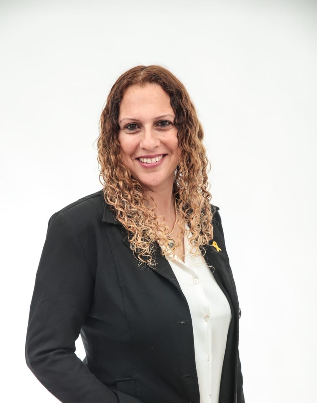 אישה במעיל שחור וחולצה שחורה יושבת ליד שולחן בידיים שלובות.