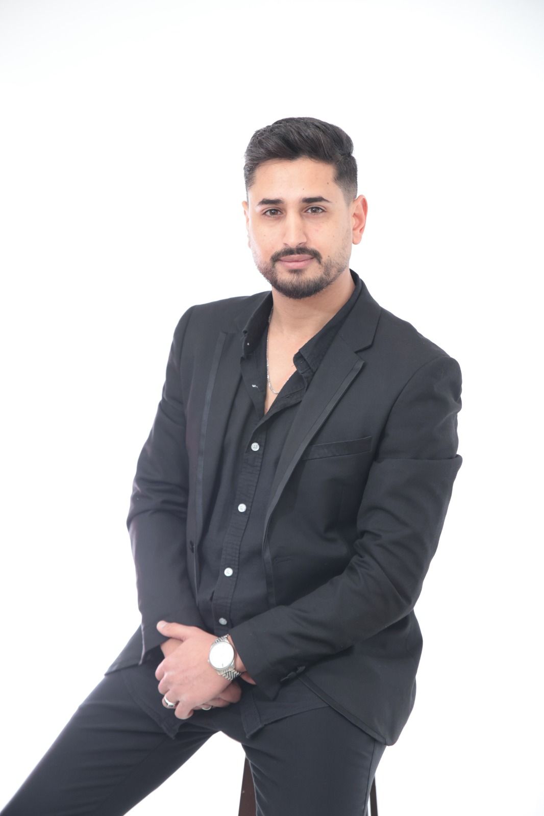 גבר בחליפה שחורה וחולצה שחורה עומד בזרועותיו שלובות.