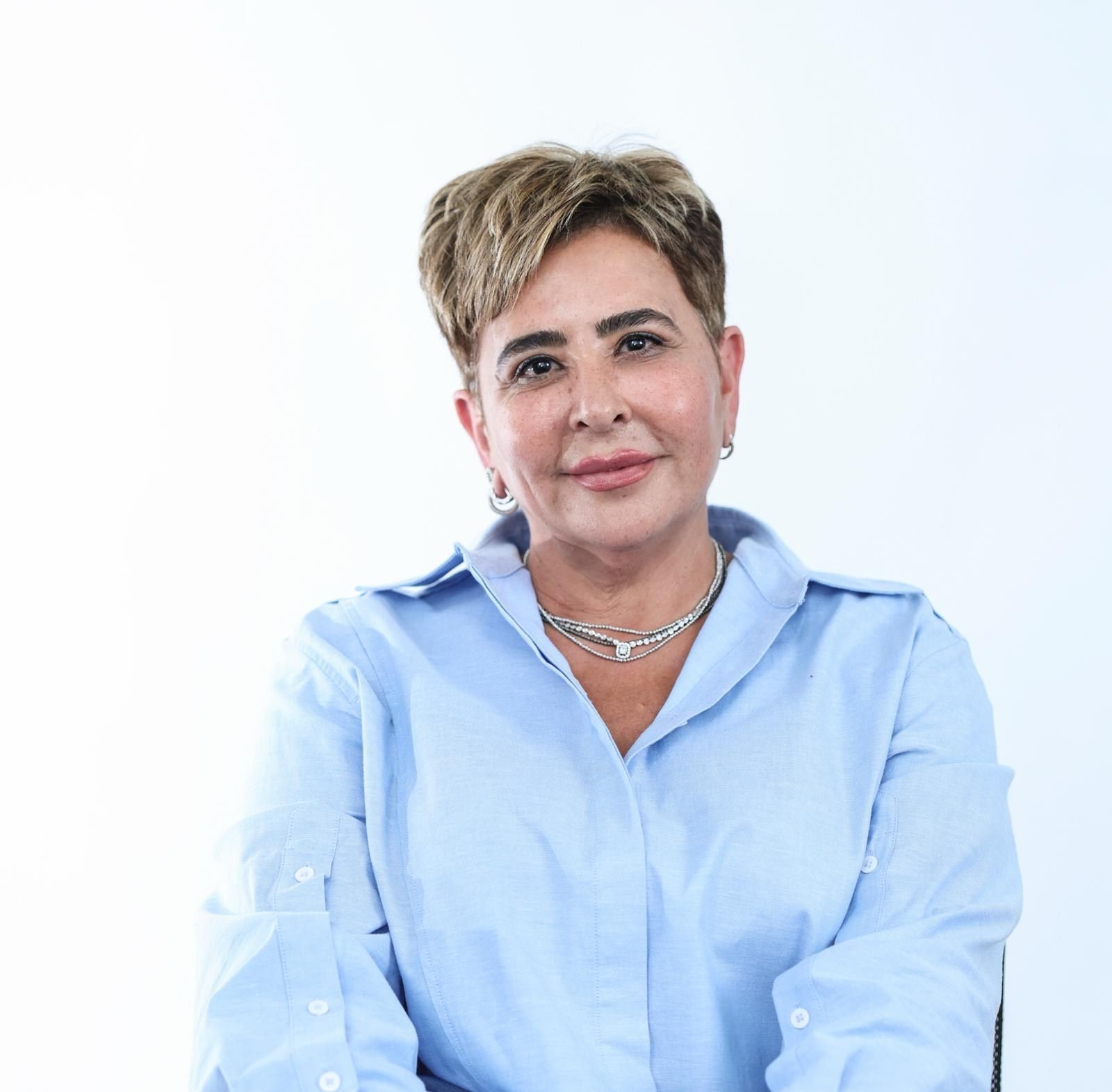 אישה במעיל שחור וחולצה שחורה יושבת ליד שולחן בידיים שלובות.