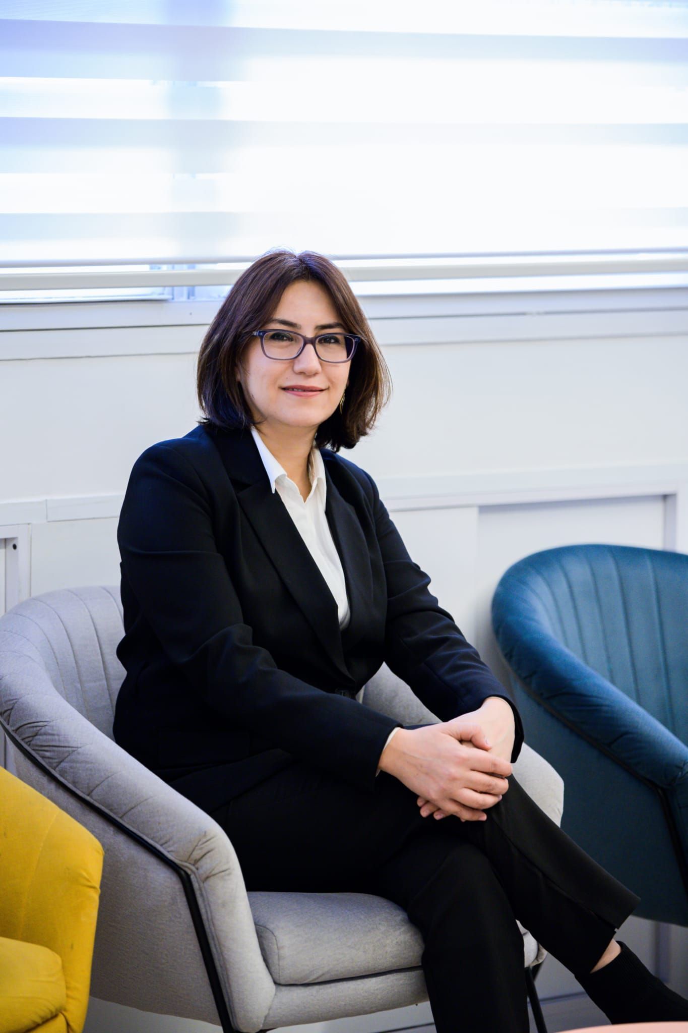 אישה מרכיבה משקפיים וז'קט שחור עומדת מול רקע לבן.