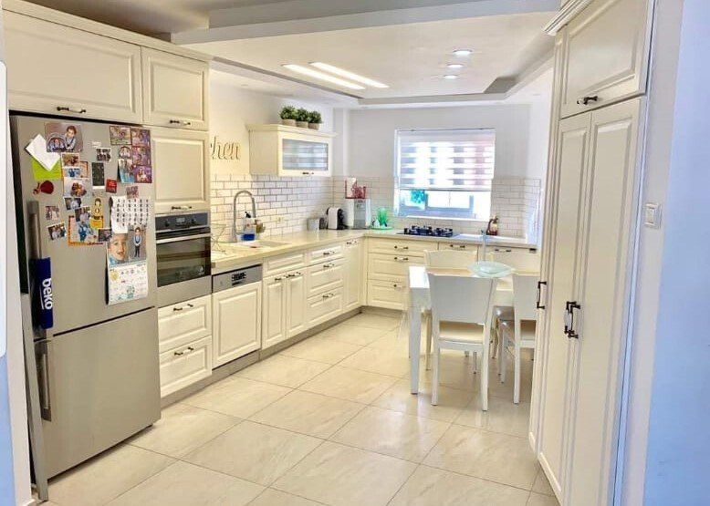 מטבח עם ארונות לבנים, מוצרי נירוסטה, מקרר, שולחן וכיסאות.