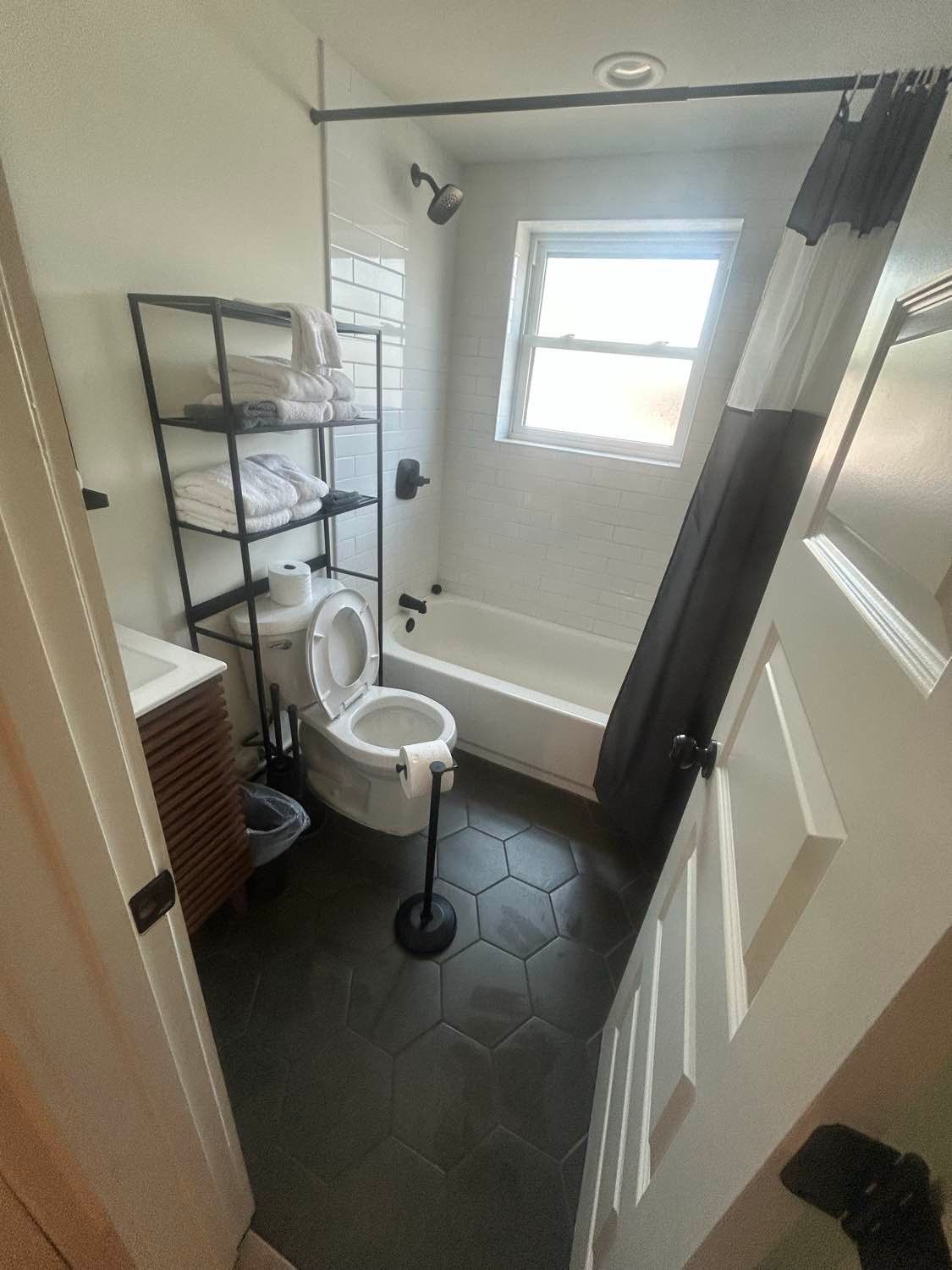 Bathroom with white walls, black hexagon tile floor, shelving, toilet, tub, and shower curtain.
