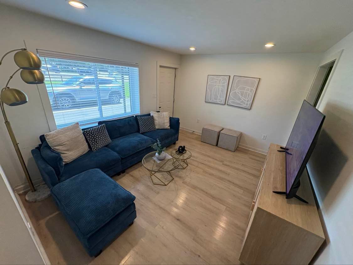 Living room with blue sofa and ottoman, light wood floor, gold lamp, and large TV.