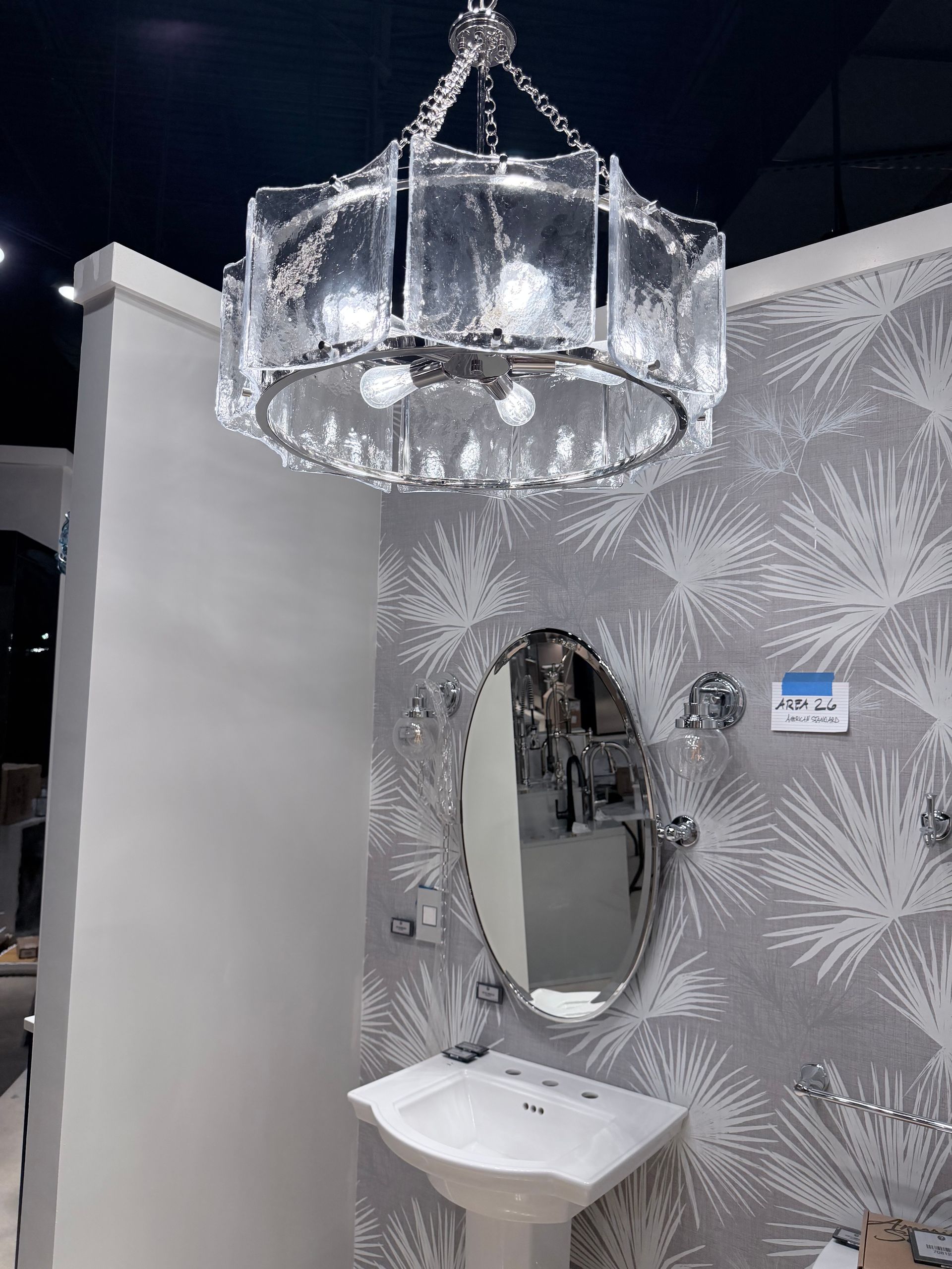 A modern bathroom vanity area with a textured glass drum chandelier, oval mirror, pedestal sink, and patterned wallpaper.