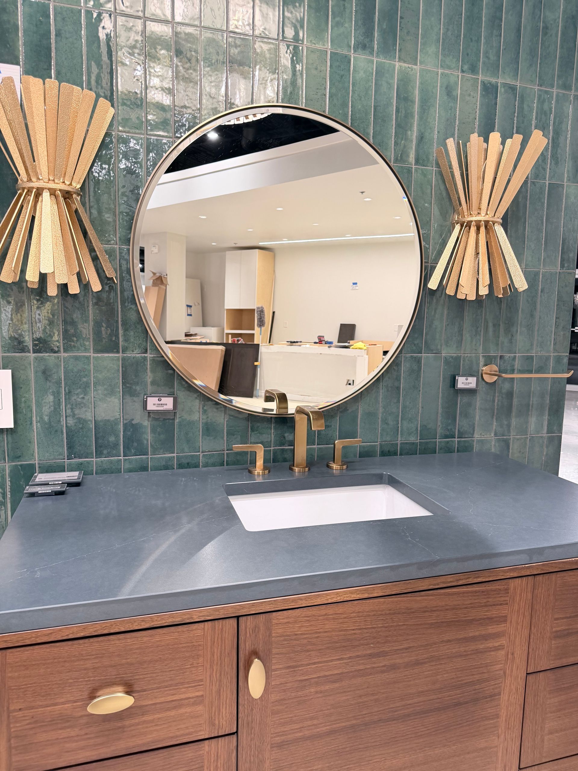 A round mirror above a wooden vanity with a grey countertop, featuring gold fixtures and green vertical wall tiles.