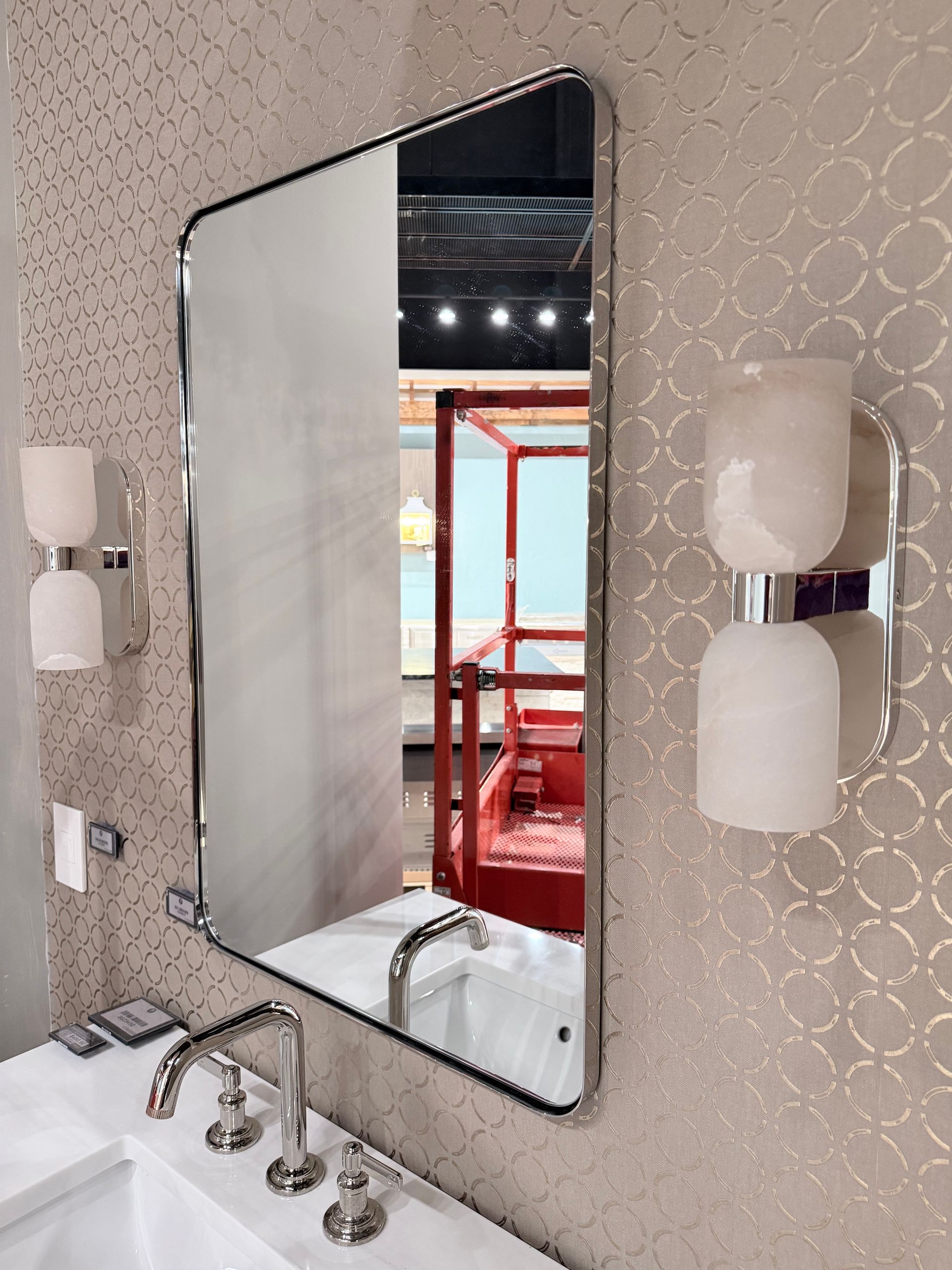 A bathroom vanity with a chrome-framed rectangular mirror, white sink, and two frosted glass wall sconces on patterned wallpaper.