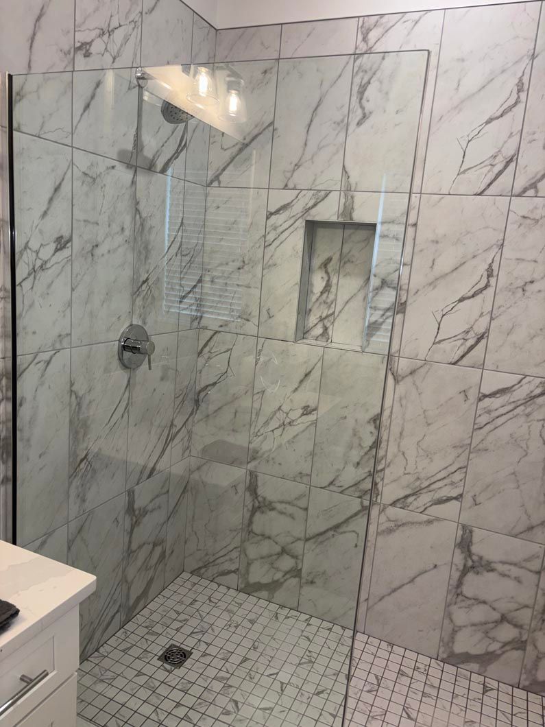 A modern walk-in shower with white and grey marble-patterned wall tiles, a built-in shelf, and a glass partition.