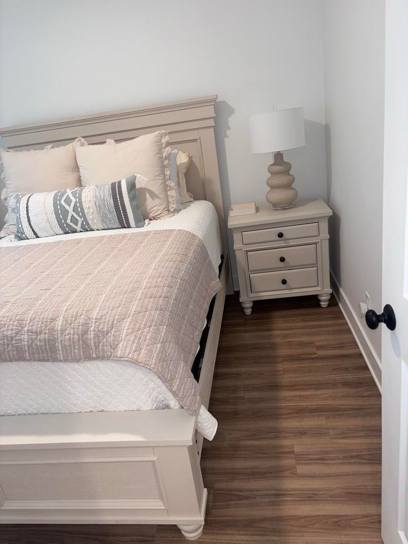A cream-colored bed with patterned pillows and a textured throw, next to a matching three-drawer nightstand with a lamp.