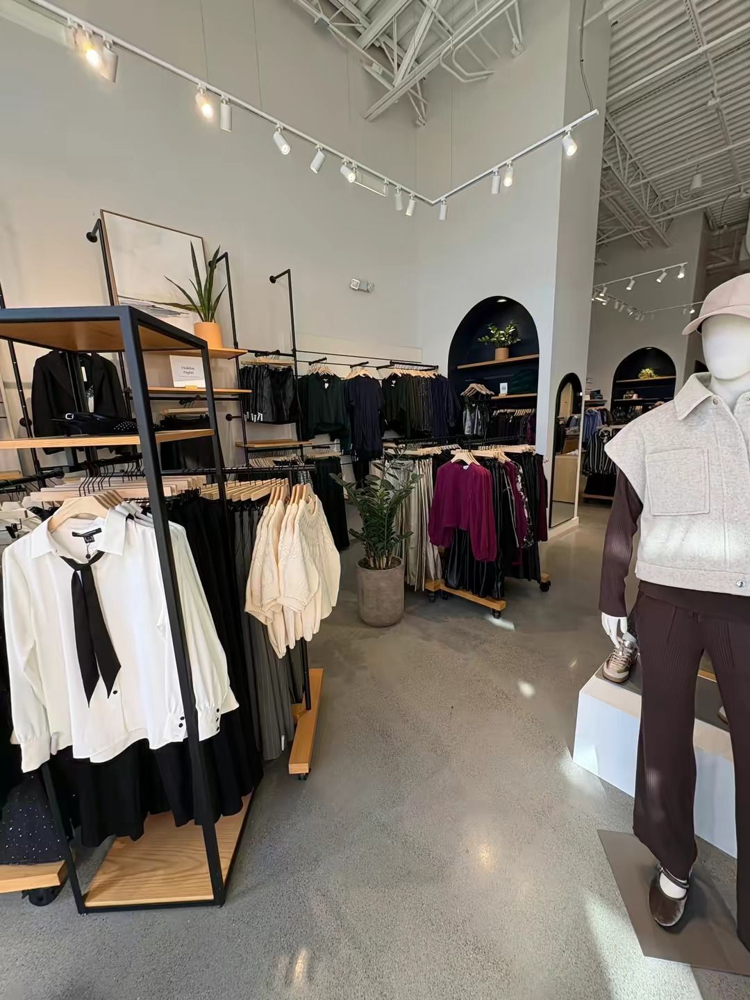 A retail clothing store interior with racks of apparel, a mannequin, potted plants, and modern lighting.