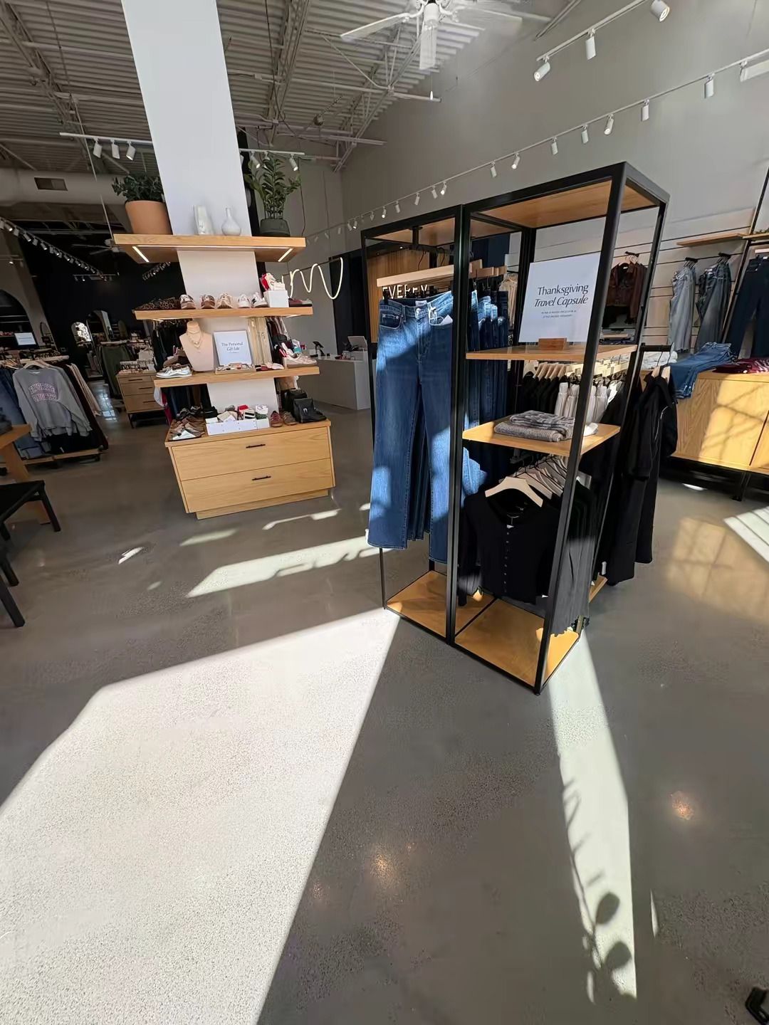 A clothing store interior featuring metal display racks of denim and shirts, and a wooden tiered shelving unit.