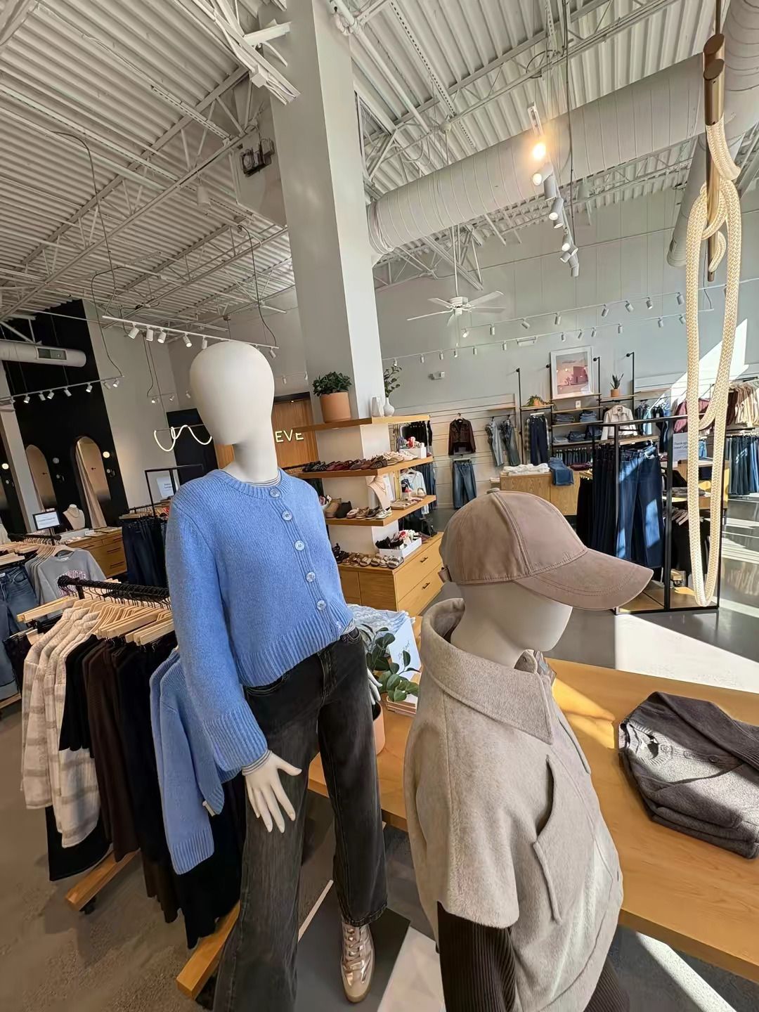 Two mannequins display a light blue cardigan and a tan baseball cap in a brightly lit clothing store.